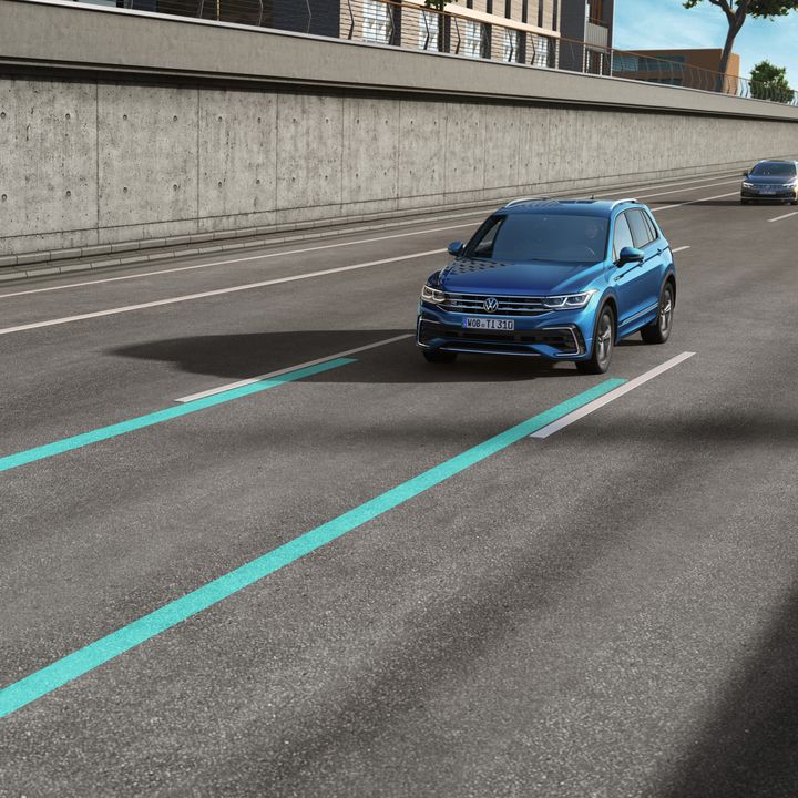 Na obrázku sú dve osobné vozidlá jazdiace po viacprúdovej ceste pri betónovej stene. Na vozovke sú vyznačené dva modré pruhy systému zmeny jazdného pruhu. Prvé auto je modré a druhé strieborné.