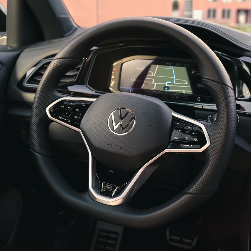 Interiér VW T-Roc, detailný pohľad na multifunkčný volant. Za volantom je digitálny prístrojový panel s displejom s navigačnou mapou. Čiastočne je vidieť otvory ventilácie, displej systému infotainment, ovládanie klimatizácie, kľučku dverí.