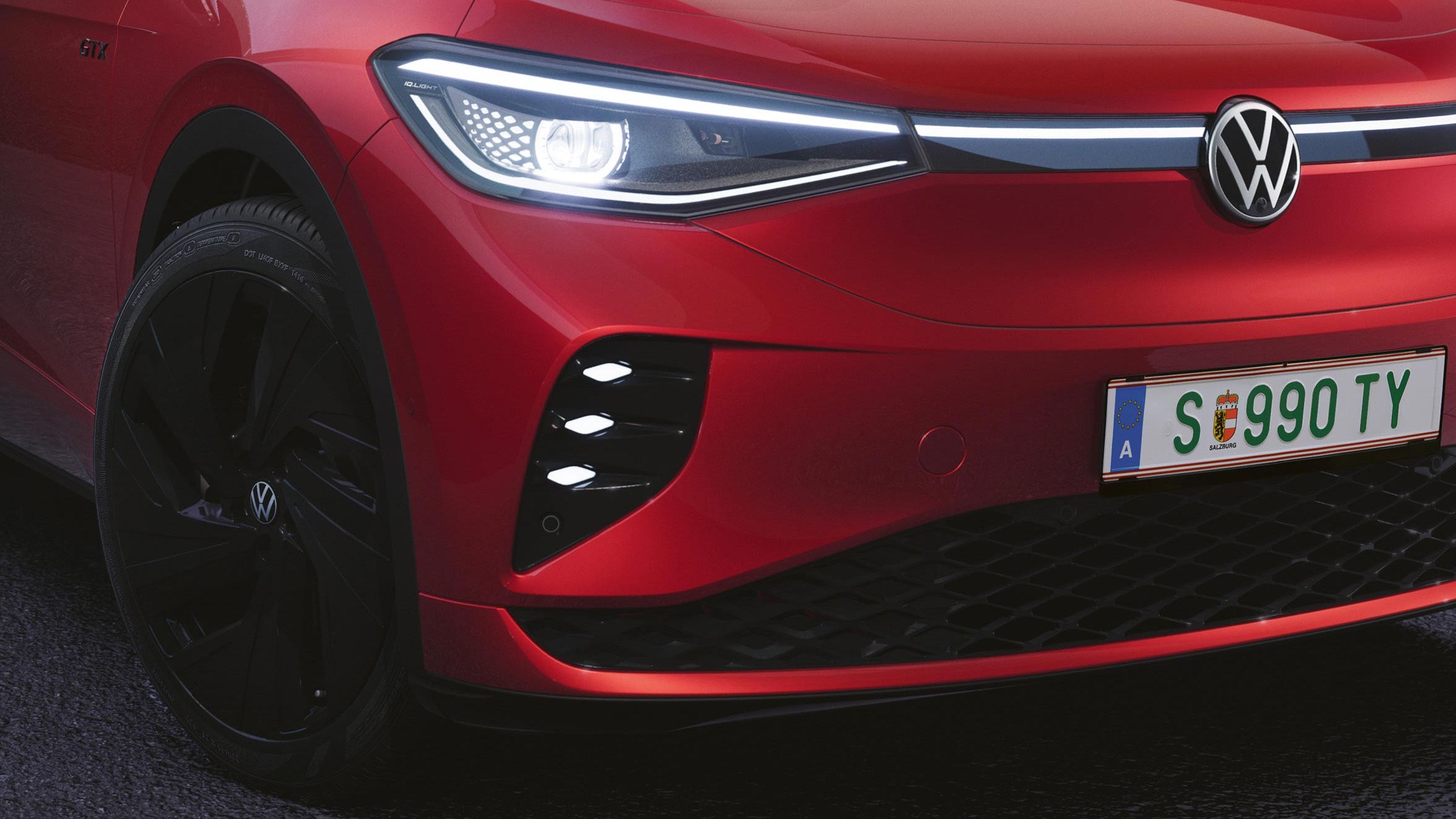 VW ID.4 GTX bočný pohľad na predné partie. Vozidlo má zapnuté moderné predné svetlomety. Červený lak vozidla dopĺňajú čierne disky kolies a čierny kryty vonkajších spätných zrkadiel.