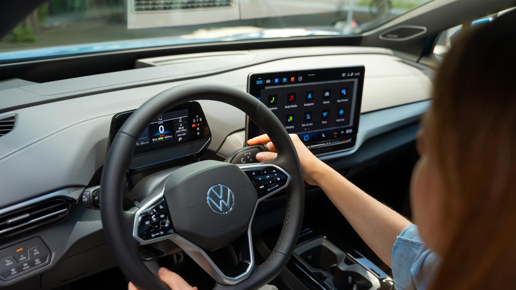 Pohľad do interiéru stojaceho vozidla VW ID.4 so ženou sediacou na mieste vodiča, ktorá pravou rukou ovláda radenie na volante.