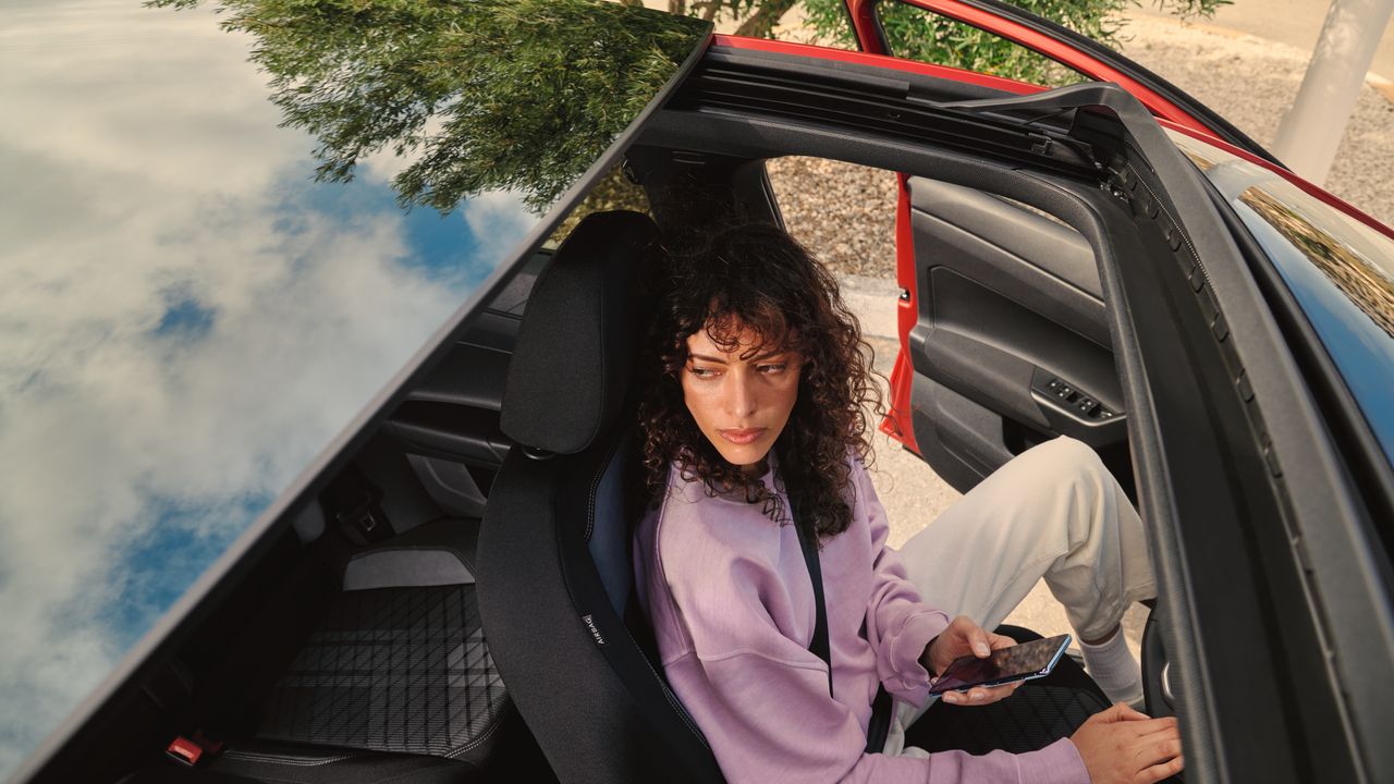 VW Taigo: Pohľad cez voliteľnú panoramatickú sklopnú/posuvnú strechu, žena s kučeravými vlasmi sedí na sedadle vodiča. Za vozidlom je strom.