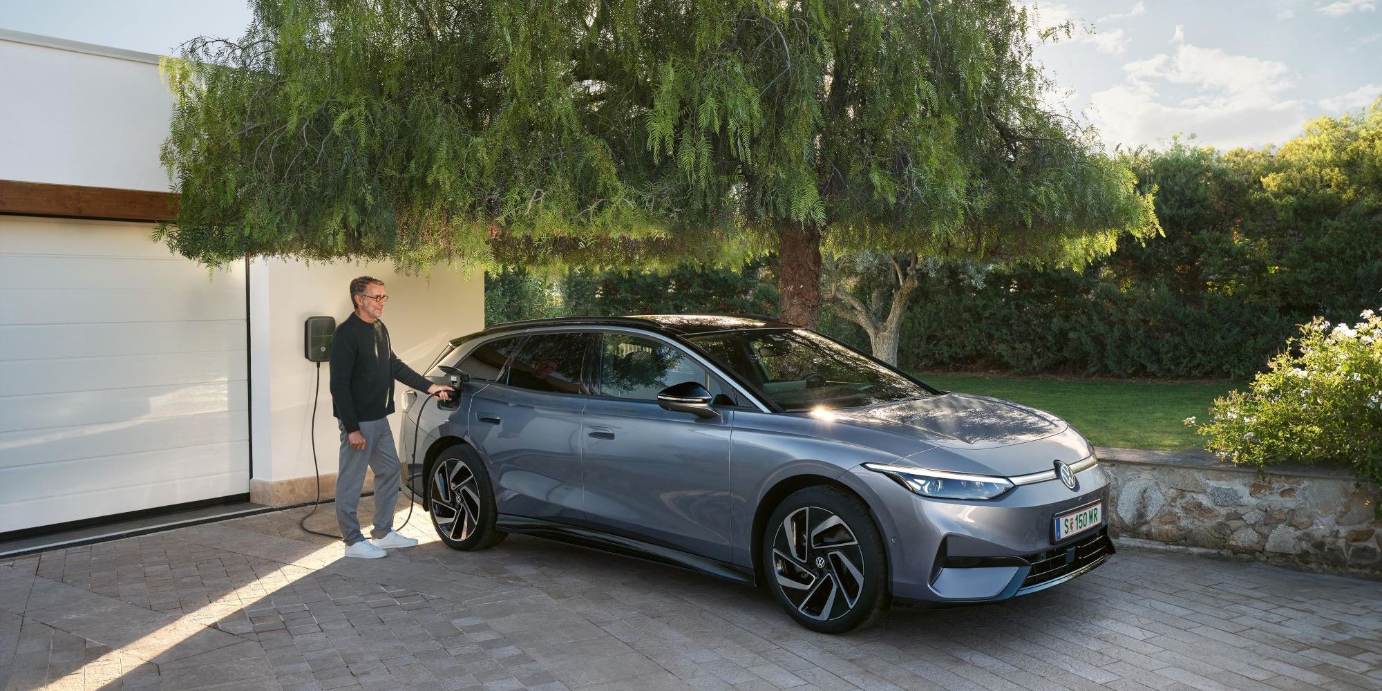 Muž stojí vedľa modrého VW ID.7 Tourer, ktorý sa nabíja pred garážou pri nabíjacej stanici Wallbox.