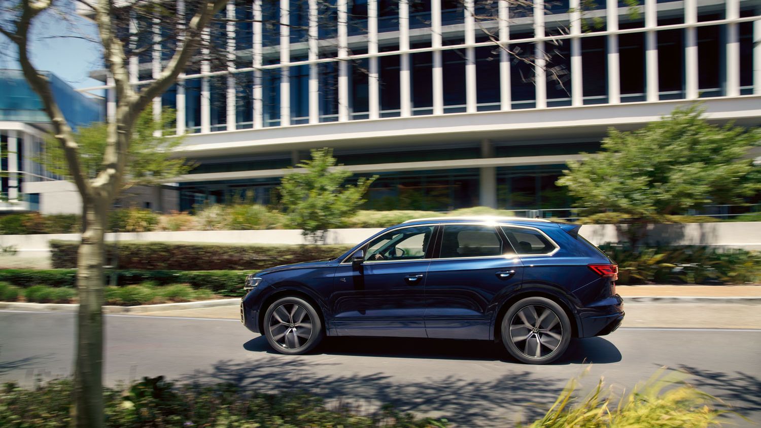 Modrý VW Touareg jazdí po ceste v meste, pričom v pozadí je moderná budova obklopená stromami a zeleňou. Slnečné svetlo sa odráža od auta.