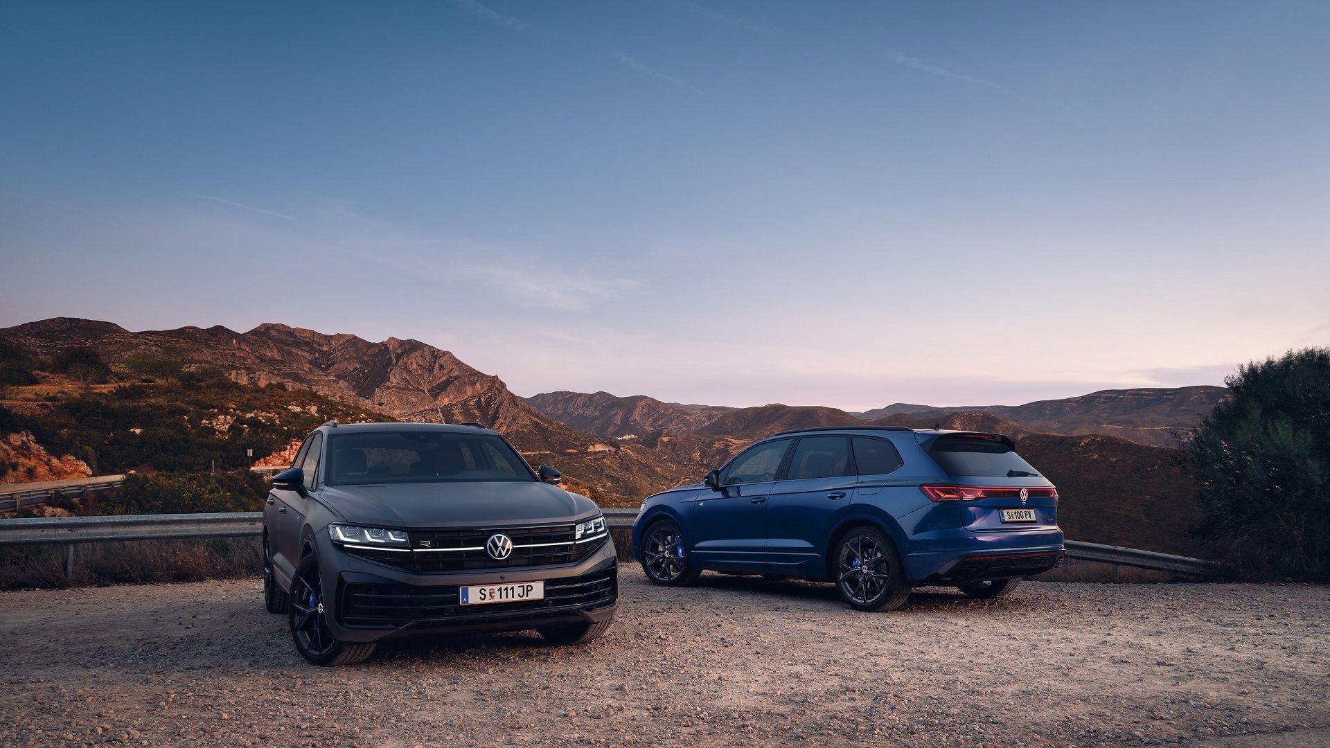 Dve vozidlá Volkswagen Touareg stoja na štrkovom priestranstve pri ceste. Z miesta je výhľad na pohorie.