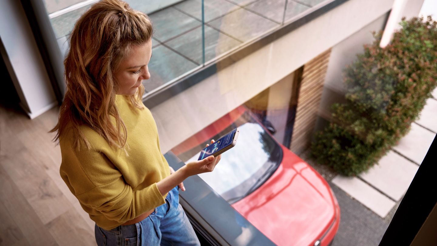 Žena sa doma prezerá svoj smartfón, zatiaľ čo jej Tiguan stojí zaparkovaný na príjazdovej ceste.