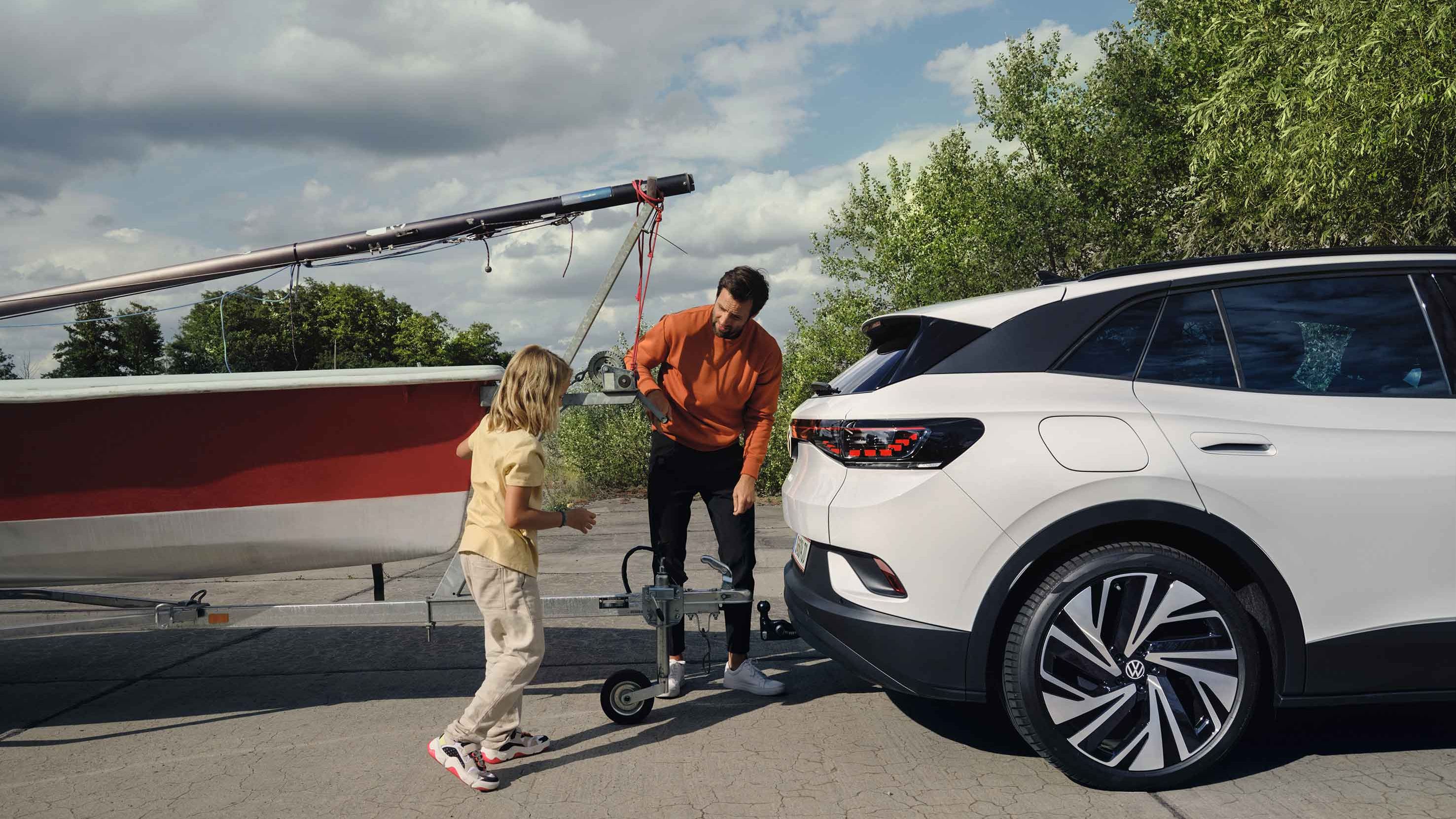 Muž a dievča zapájajú prívesný vozík s loďou k ťažnému zariadeniu elektrického SUV. Okolo nich je zeleň s niekoľkými stromami a čiastočne zamračenou oblohou.