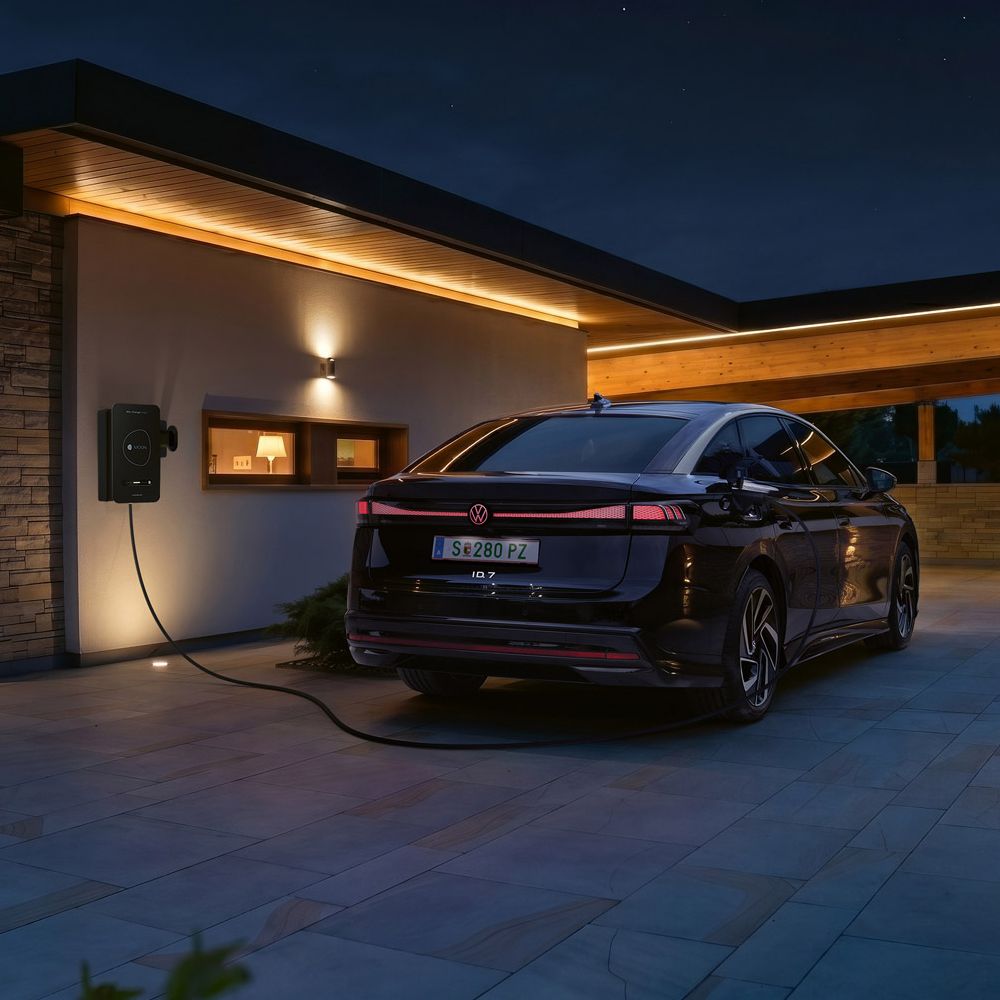 Čierne elektrické auto je zaparkované na príjazdovej ceste moderného domu v noci a je pripojené k domácej nabíjacej stanici. Teplé, nepriame svetlo na fasáde a prístrešku osvetľuje scénu. V pozadí sa črtajú kopce za súmraku.
