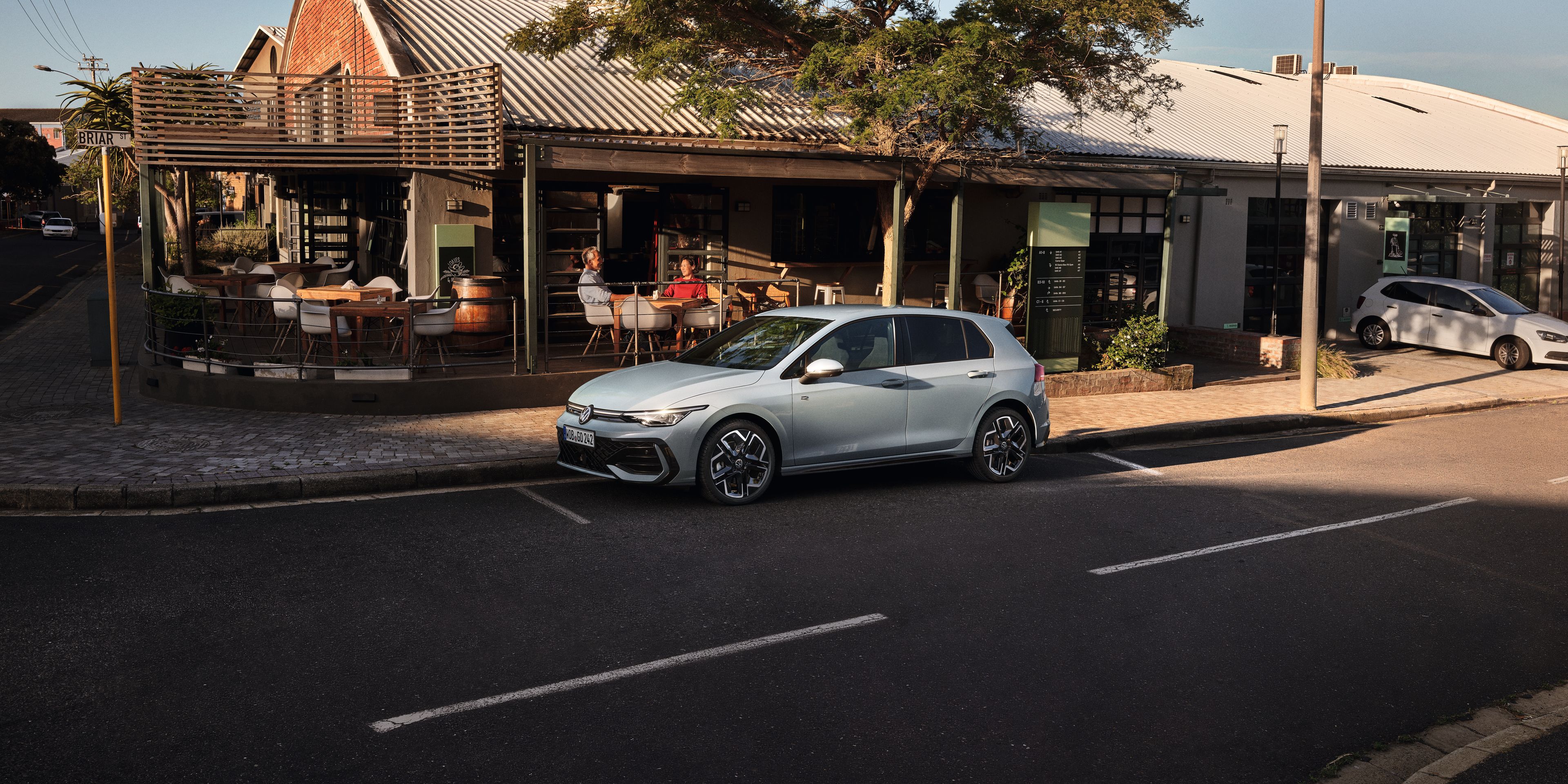 Biely Golf R-Line zaparkovaný na parkovacom mieste na ceste pred reštauráciou. Za vozidlom je reštaurácia a na terase sedia zákazníci.