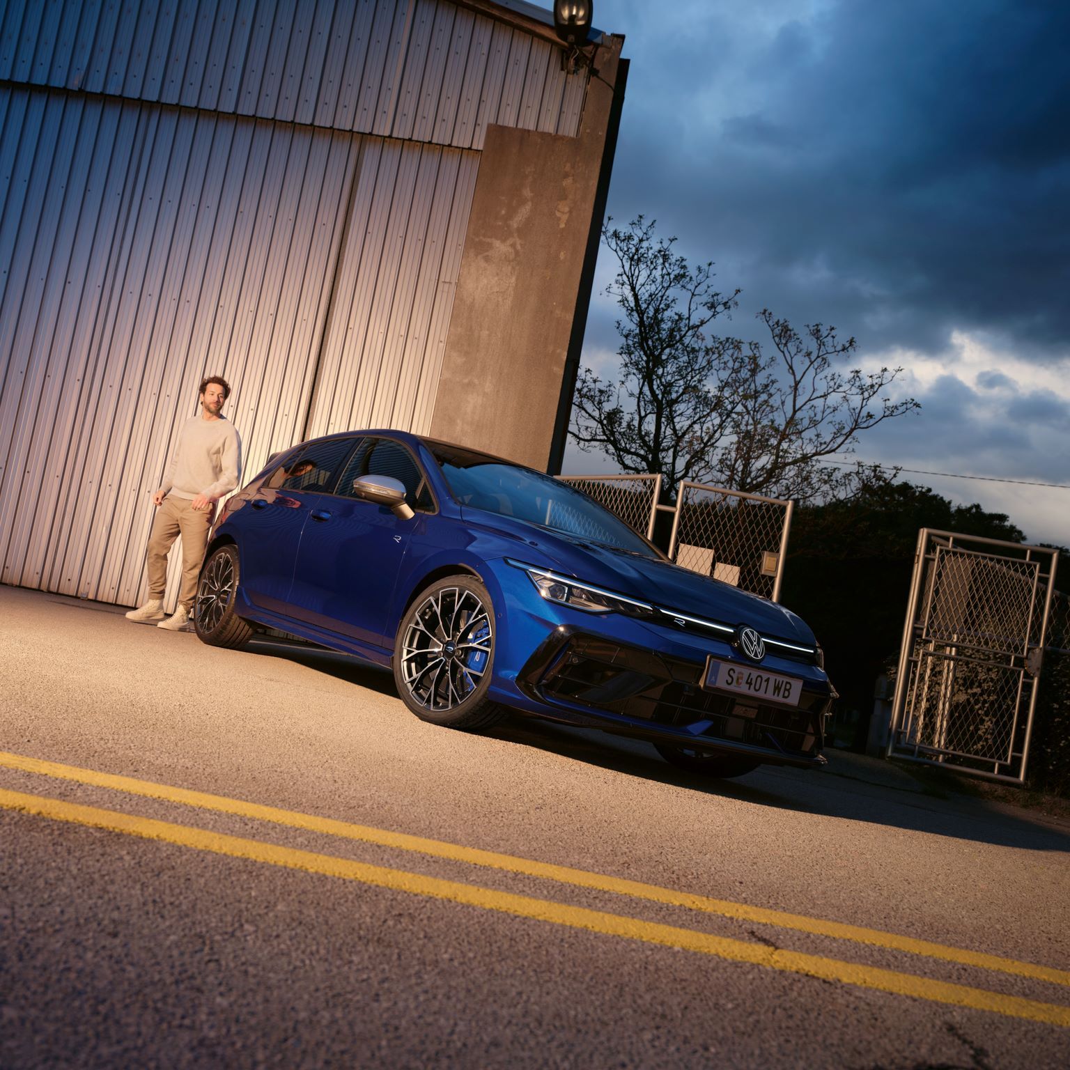 Modrý VW Golf R zaparkovaný pred industriálnou budovou a stojí pri ňom muž. Obloha je zatiahnutá, je šero a svieti pouličná lampa.