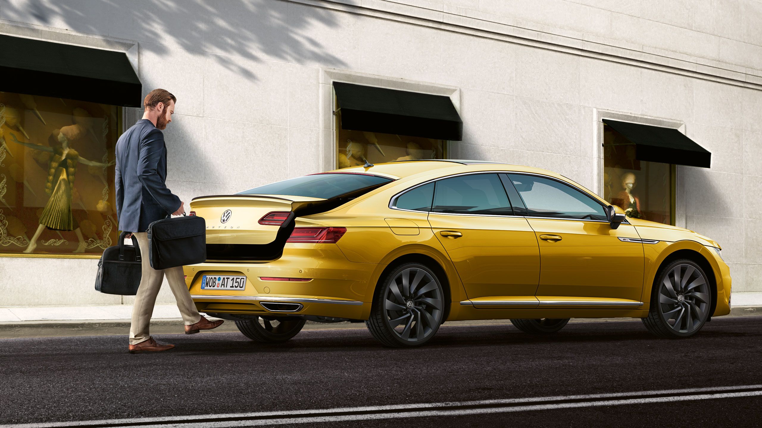 Na obrázku je elegantne oblečený muž, ktorý otvára batožinový priestor pohybom nohy pod zadným nárazníkom a kladie čierne kufre dovnútra automobilu Volkswagen zaparkovaného pred budovou s výkladmi.