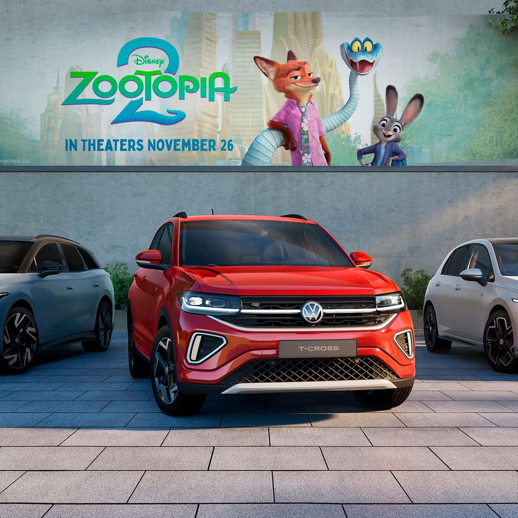 Tri modely automobilov Volkswagen (ID.4X, T-Cross a Golf) zaparkované pred billboardom s filmom Zootopia 2. V pozadí sú stromy a zeleň. Automobily sú rôznych farieb: modrá, červená a biela.