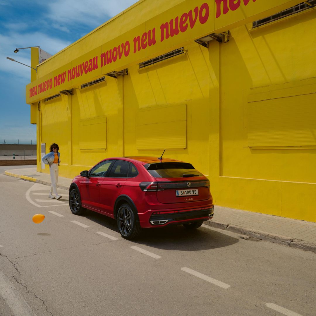 Červené Taigo R-Line je zaparkované pred žltou budovou s nápisom "neuvo neu nouveau neu nuevo neu". K vozidlu sa popri stene približuje mladá žena v modrej bunde. Vozidlo stojí na parkovacom mieste pri ceste.