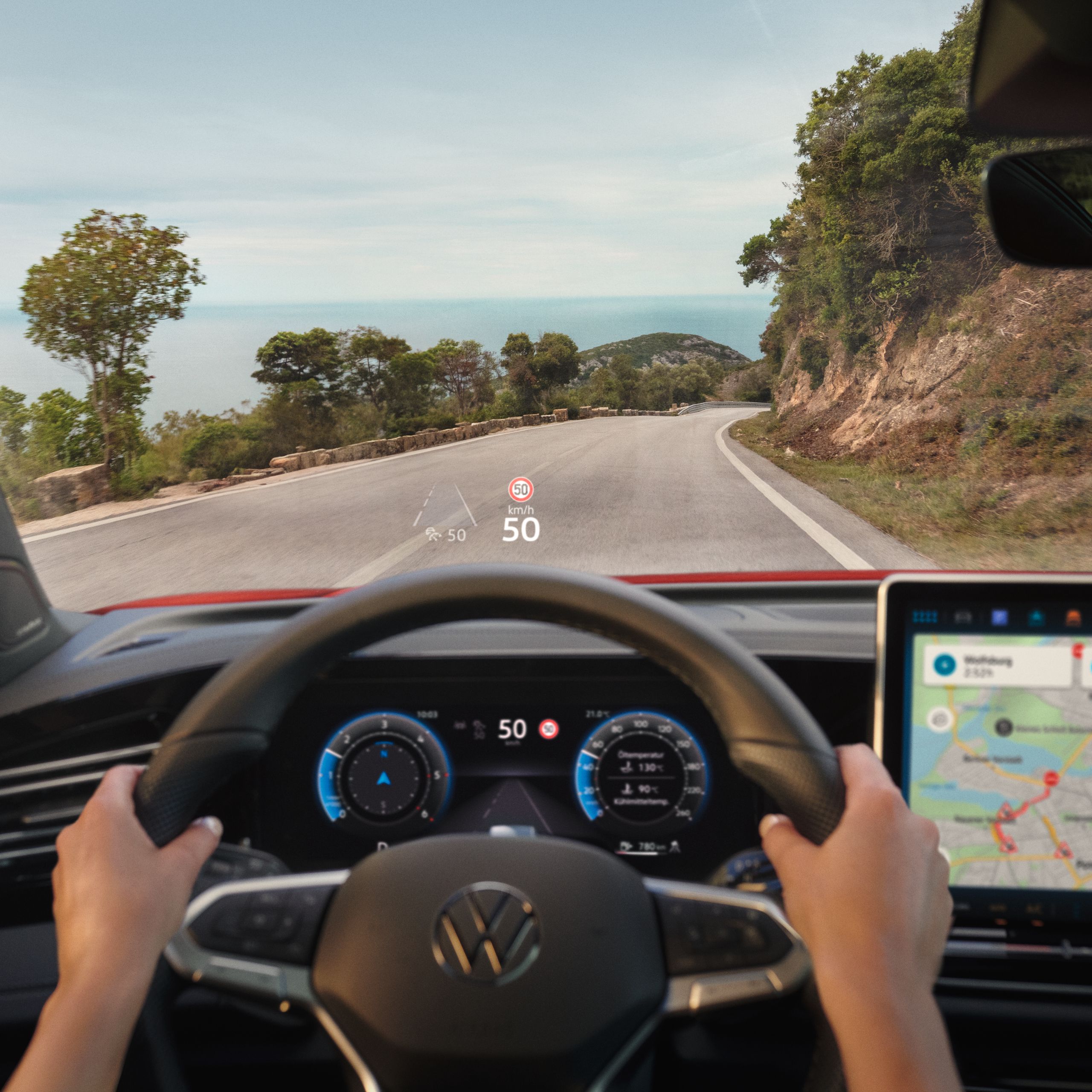 Pohľad do interiéru VW Golf. Za volantom sedí osoba a riadi vozidlo. Vidíme tiež digitálne prístroje, ventiláciu, displej systému infotainment so spustenou navigáciou. Na čelnom skle je zreteľný head-up displej a cez okno vidieť cestu v horách.