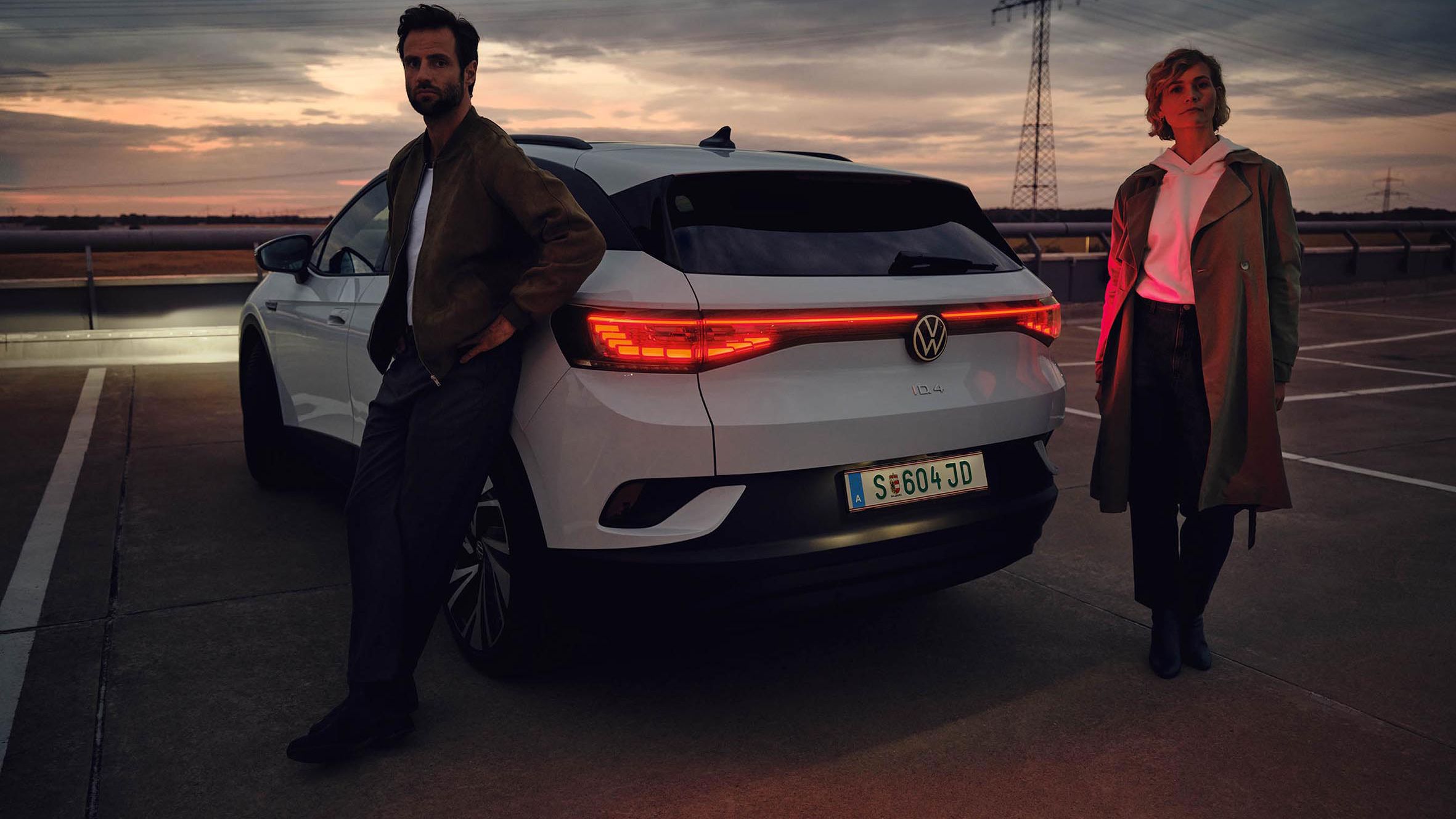 Žena a muž stoja pri bielom aute Volkswagen na parkovisku pri západe slnka. Muž sa opiera o auto, žena stojí vedľa auta. Vozidlo má zapnuté svetlá. V pozadí sú stožiare elektrického vedenia.