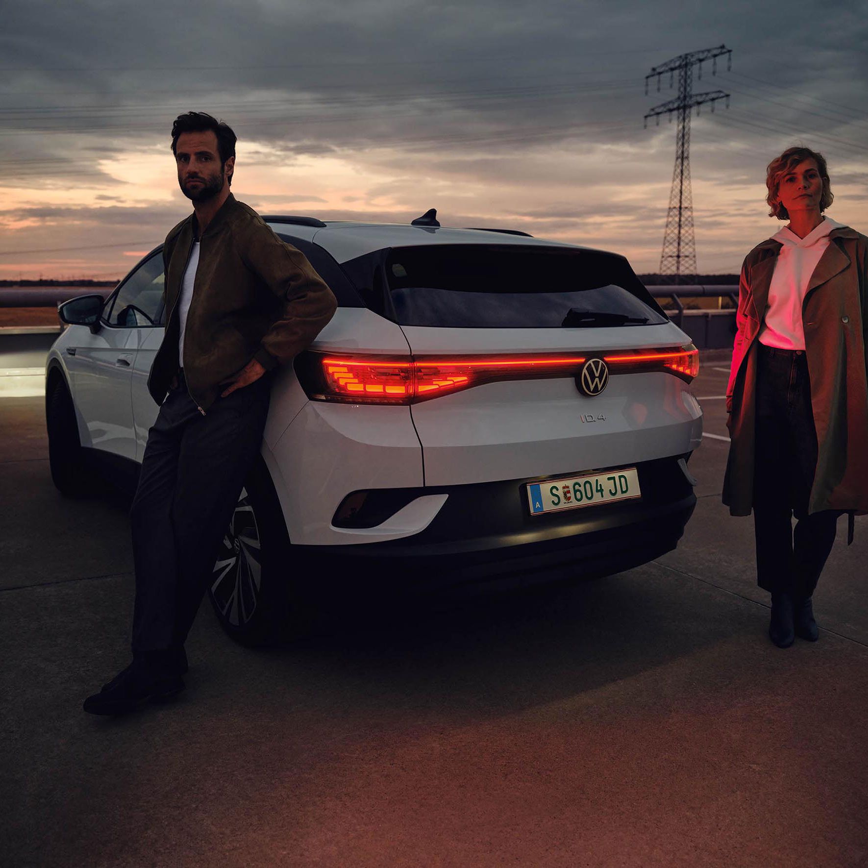 Žena a muž stoja pri bielom aute Volkswagen na parkovisku pri západe slnka. Muž sa opiera o auto, žena stojí vedľa auta. Vozidlo má zapnuté svetlá. V pozadí sú stožiare elektrického vedenia.