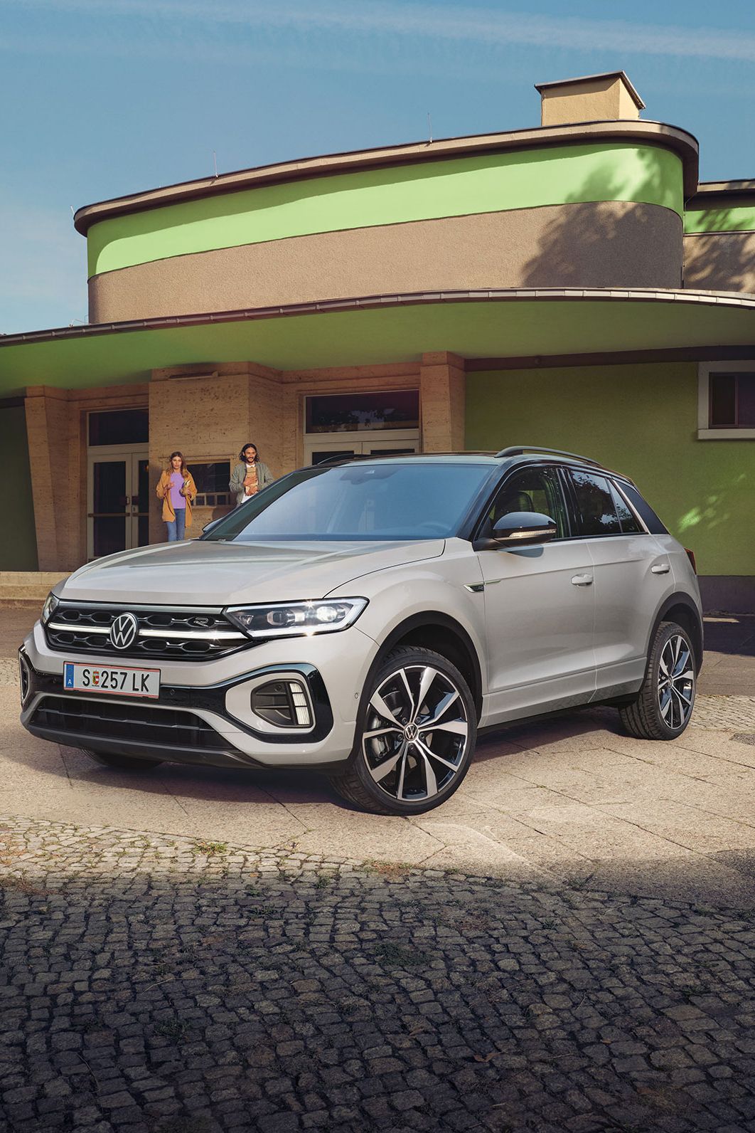 Strieborné SUV Volkswagen T-Roc je zaparkované na ulici pred budovou so svetlohnedou, zelenou fasádou a zaoblenými tvarmi. Budova je obklopená zeleňou. 