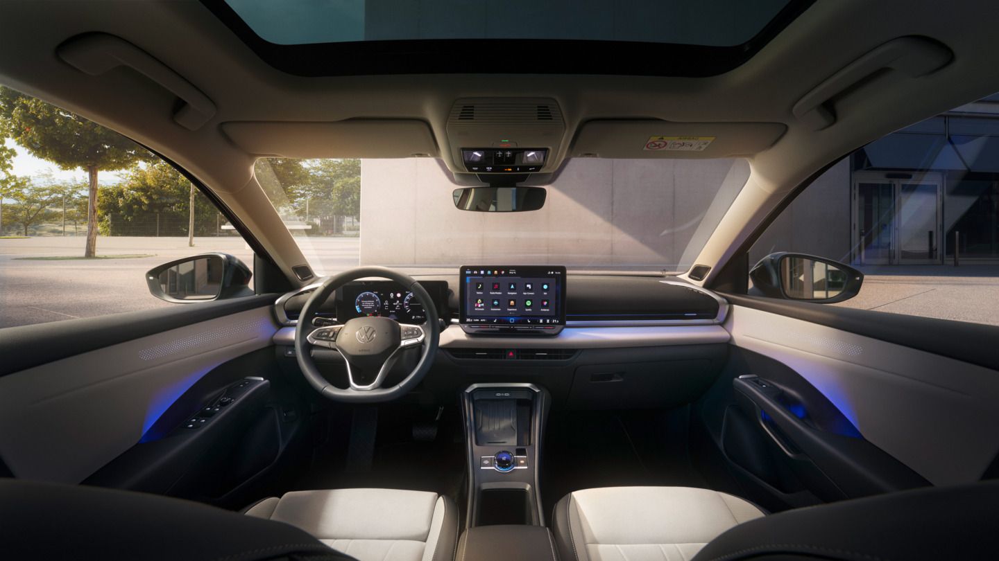 Obrázok zobrazuje interiér vozidla T-Roc so slnečnou alebo sklenenou strechou. Kokpit má veľký displej a volant s viacerými funkciami. Von je vidieť park a budovu.