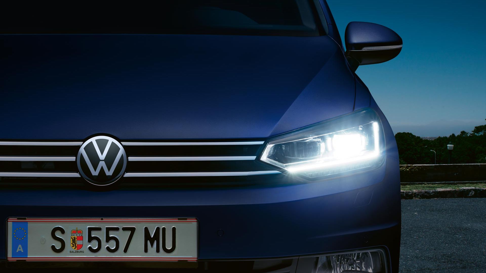 Modré auto značky Volkswagen zaparkované von za súmraku. So zasvietenými svetlometmi. Za vozidlom je zeleň a mraky v pozadí.