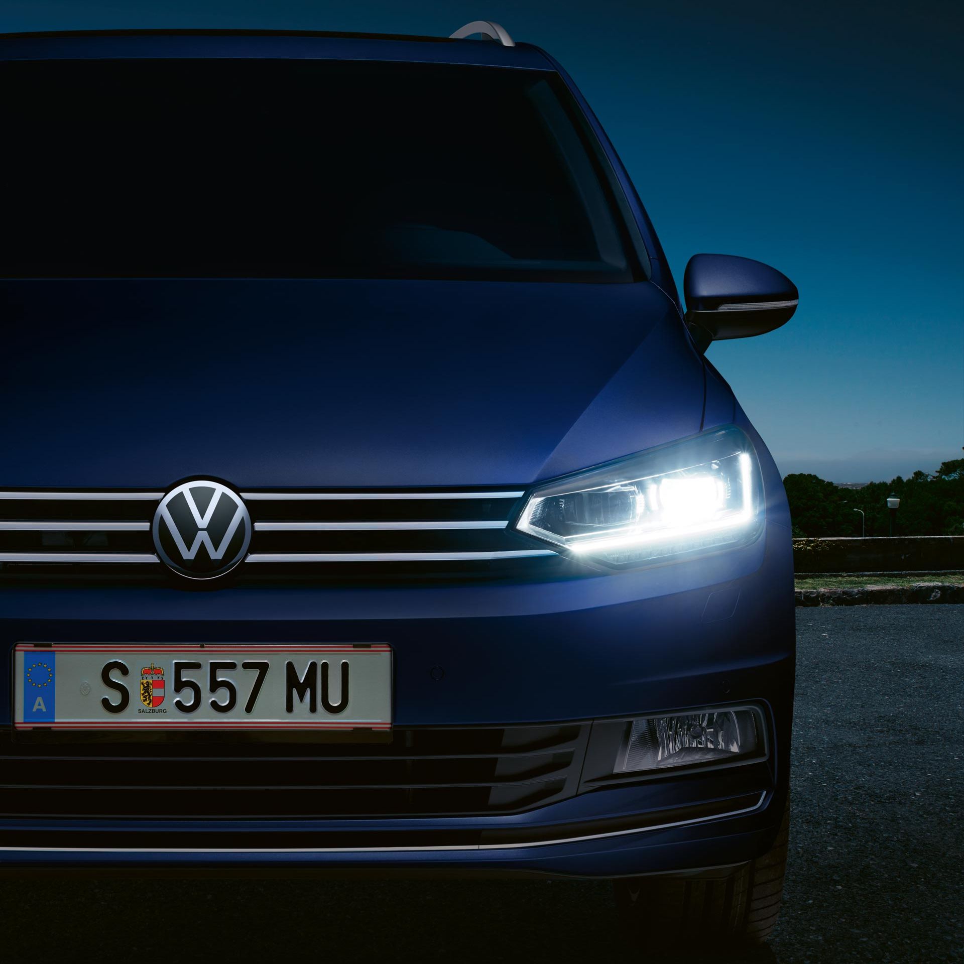 Modré auto značky Volkswagen zaparkované von za súmraku. So zasvietenými svetlometmi. Za vozidlom je zeleň a mraky v pozadí.
