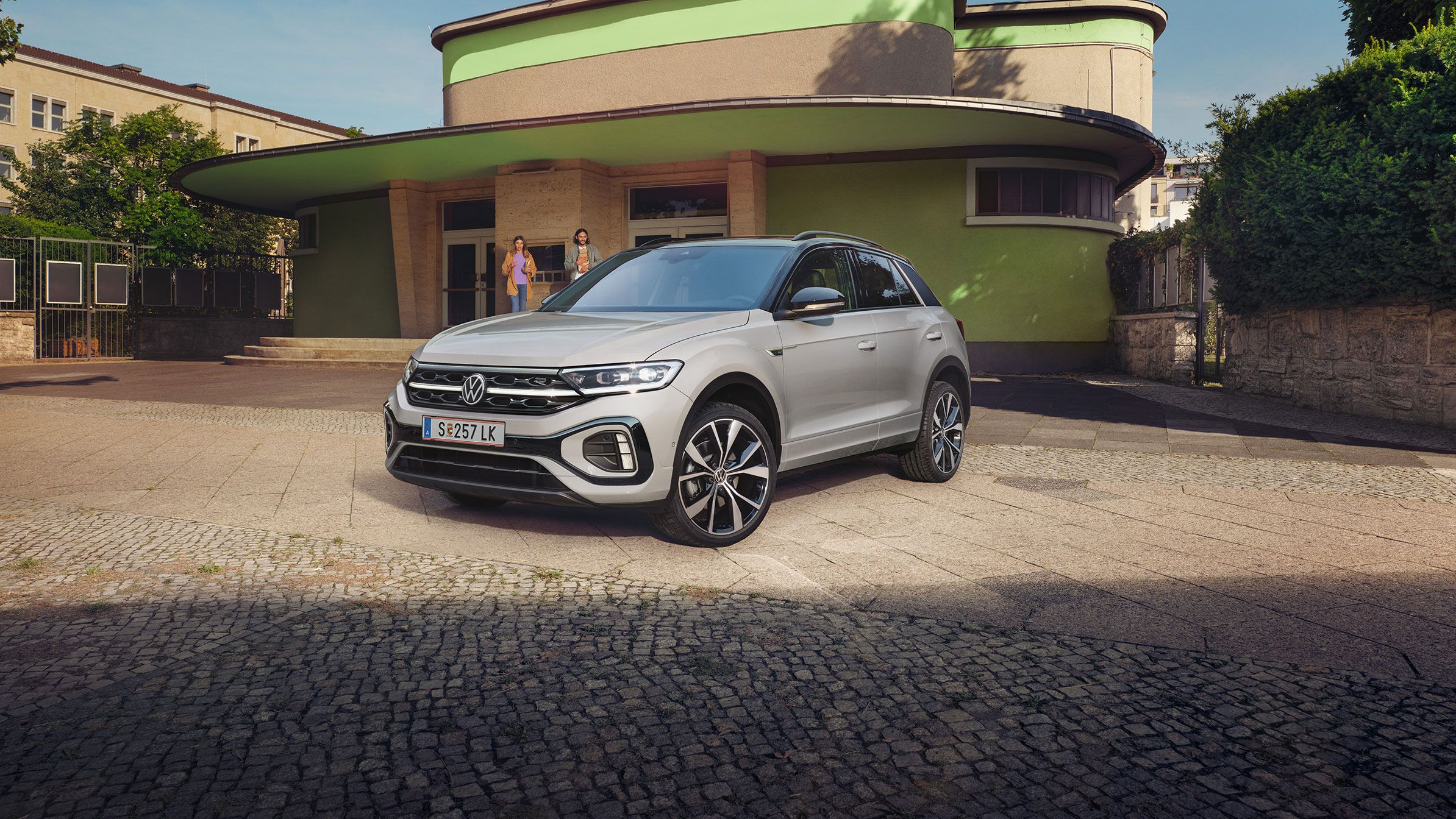 Strieborné SUV Volkswagen T-Roc je zaparkované na ulici pred budovou so svetlohnedou, zelenou fasádou a zaoblenými tvarmi. Budova je obklopená zeleňou. 