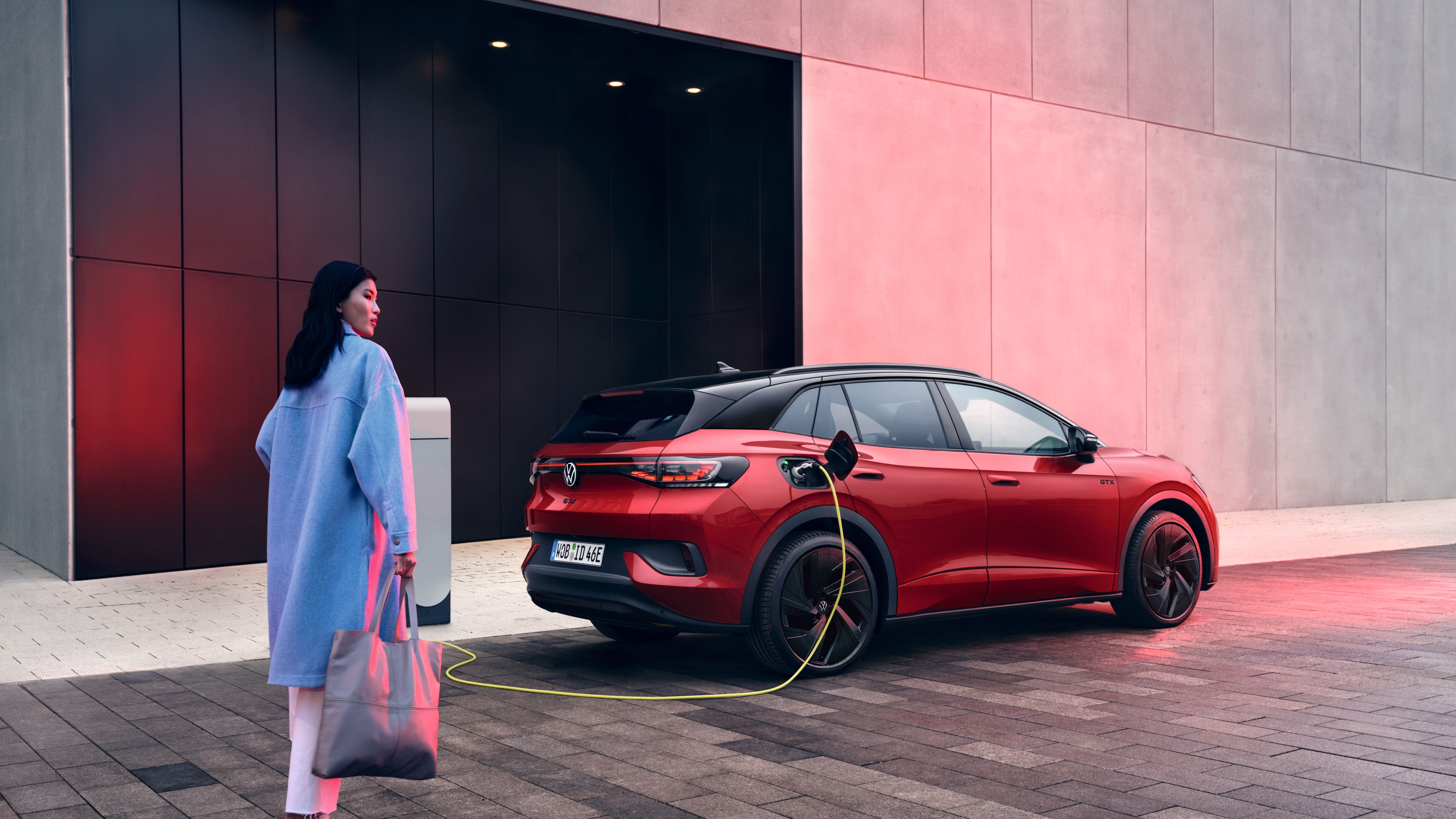 Žena stojí vedľa červeného elektrického SUV ID.4 GTX, ktoré sa nabíja pri modernej stavbe. SUV sa nabíja pri nabíjacej stanici vedľa budovy. Žena drží sivú tašku, je oblečená v modrom kabáte a ide smerom k aute.