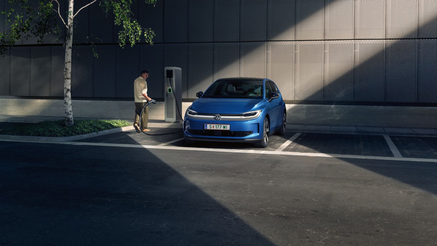 Modré ID. Polo stojí na parkovisku vedľa nabíjacej stanice. V pozadí je vidieť budovu a strom.