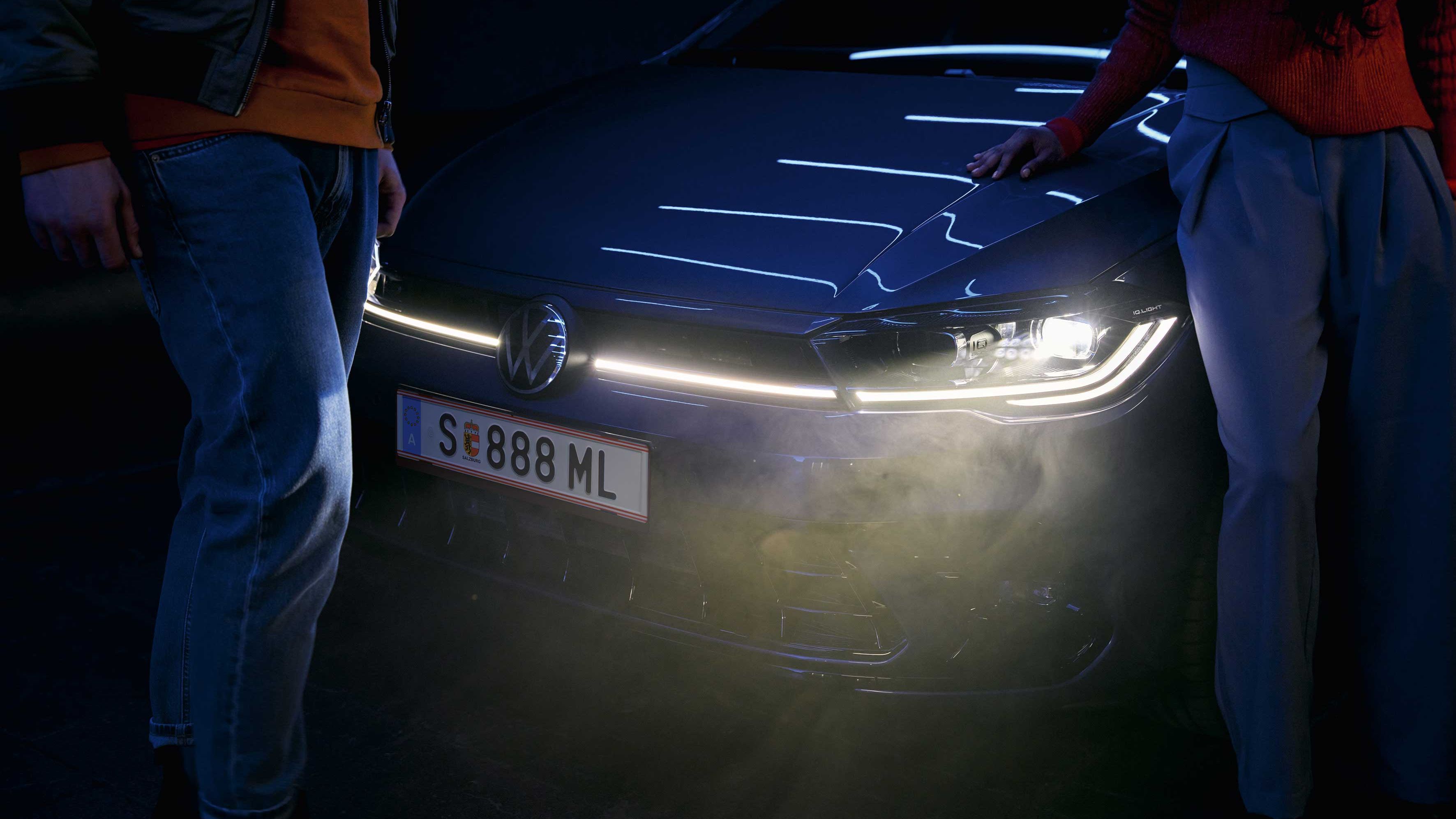 Dvaja ľudia stoja pred moderným automobilom Volkswagen so zapnutými svetlometmi v noci. Jeden z nich sa opiera rukou o kapotu auta. 