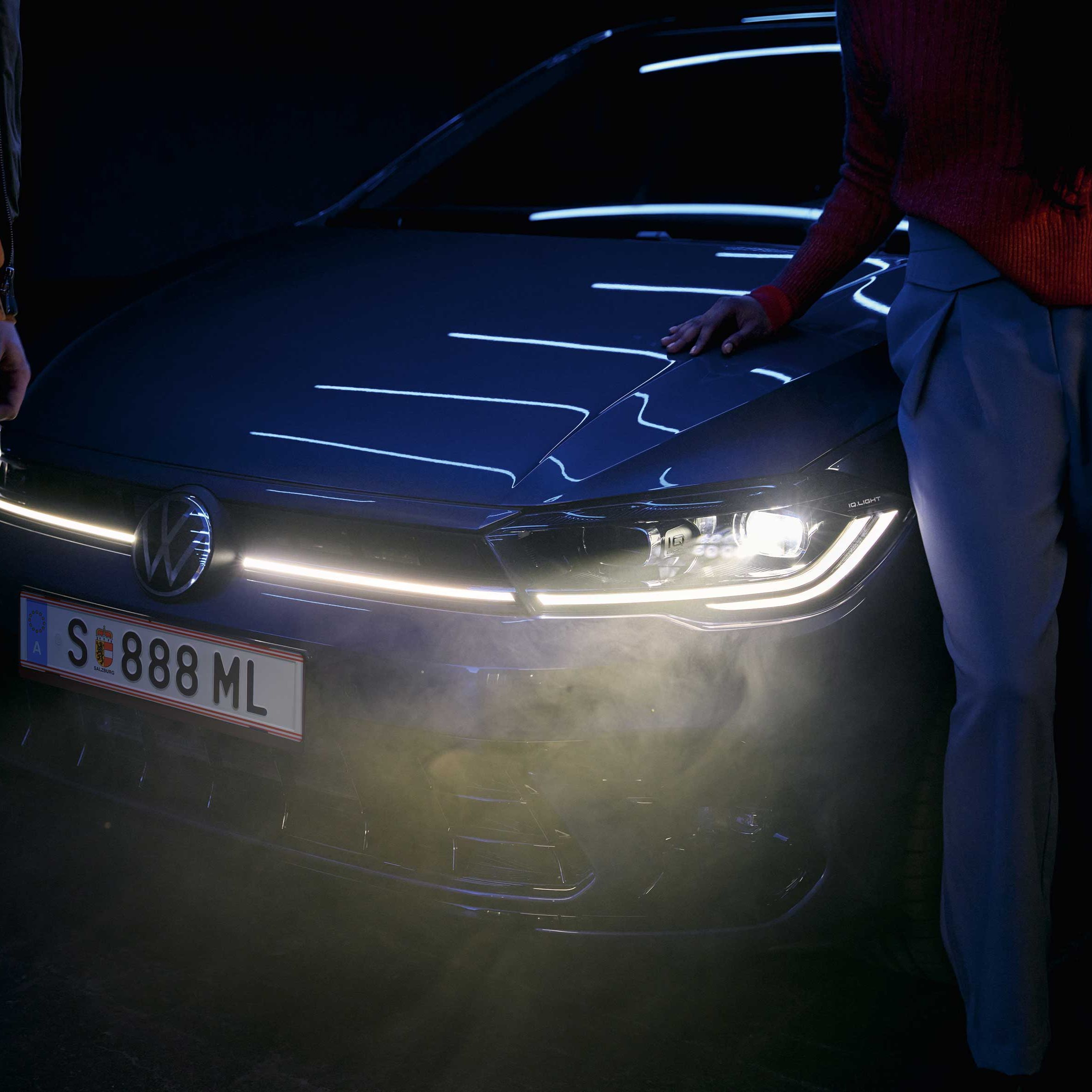 Dvaja ľudia stoja pred moderným automobilom Volkswagen so zapnutými svetlometmi v noci. Jeden z nich sa opiera rukou o kapotu auta. 