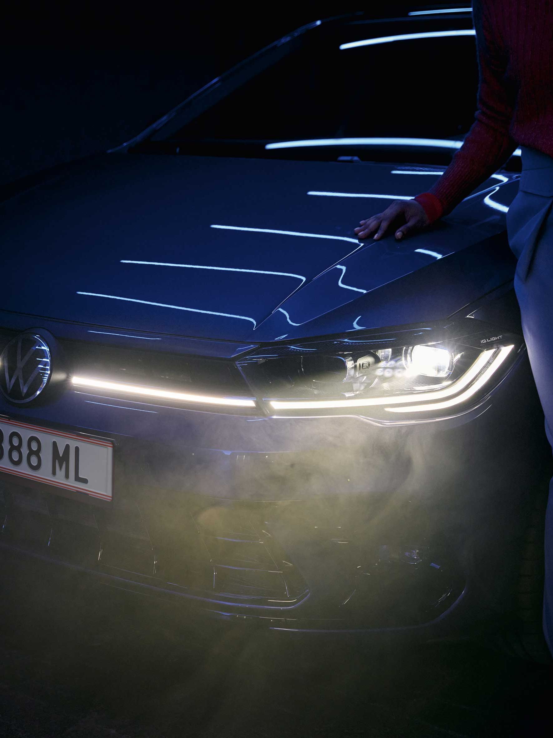 Dvaja ľudia stoja pred moderným automobilom Volkswagen so zapnutými svetlometmi v noci. Jeden z nich sa opiera rukou o kapotu auta. 