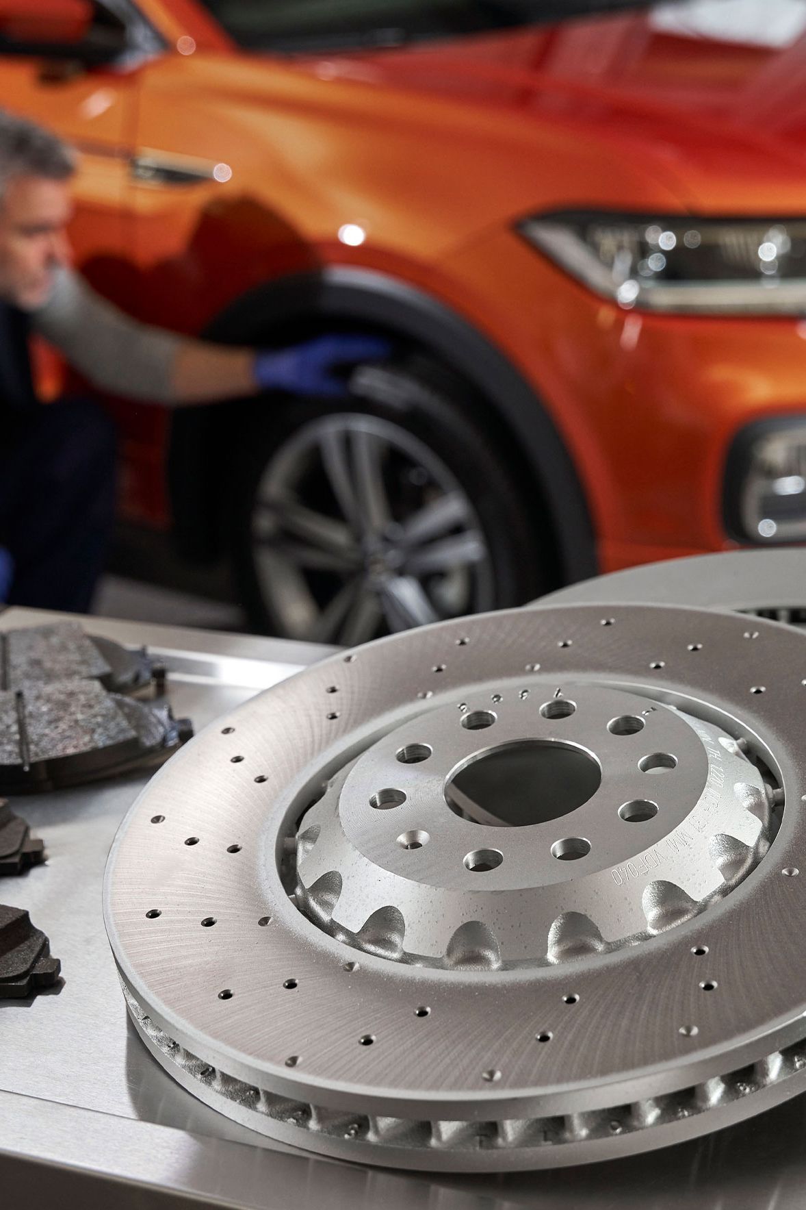 Mechanik kontroluje oranžové SUV, zatiaľ čo na stolíku sú umiestnené nové brzdové kotúče a brzdové doštičky.