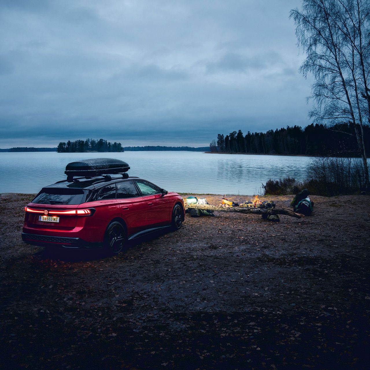 Volkswagen ID.7 Tourer GTX je zaparkovaný na brehu jazera a má zasvietené svetlá. Pred vozidlom vidieť oheň a veci na kempovanie.