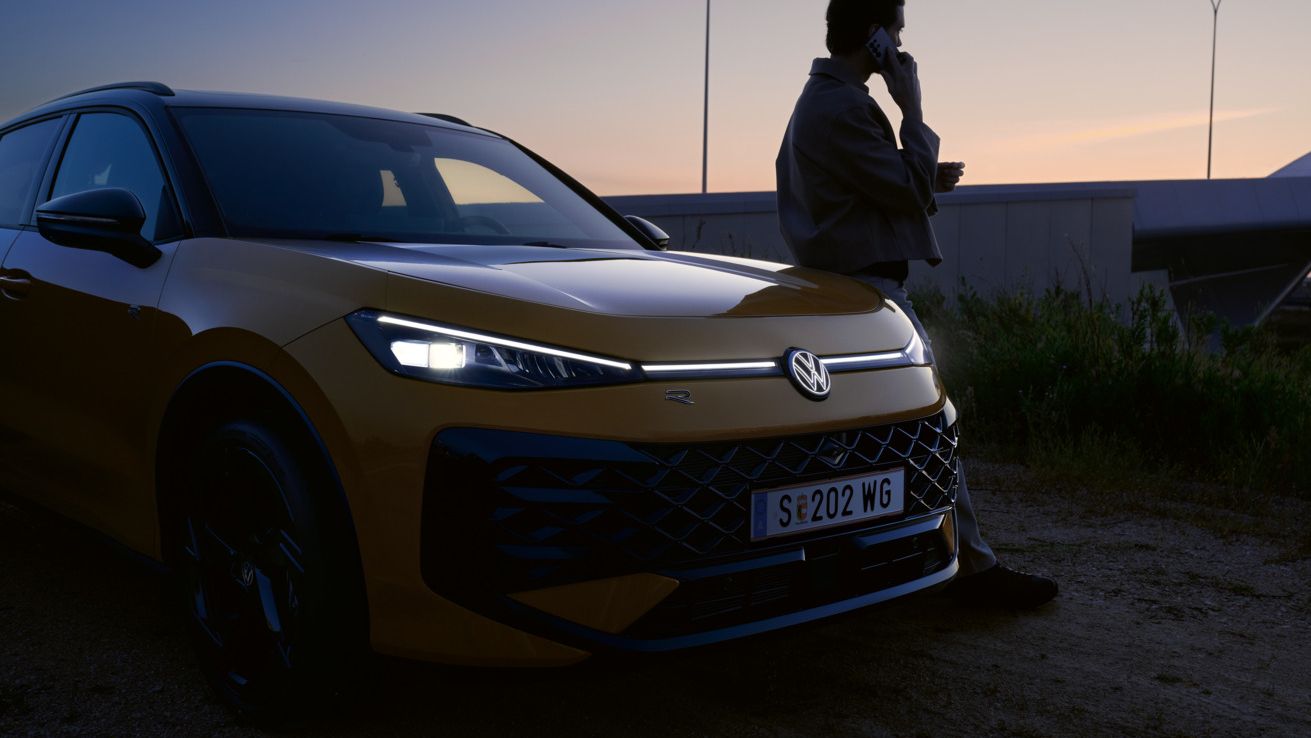 Muž sa opiera o vozidlo VW T-Roc R-Line so zapnutými svetlami a osvetleným logom.