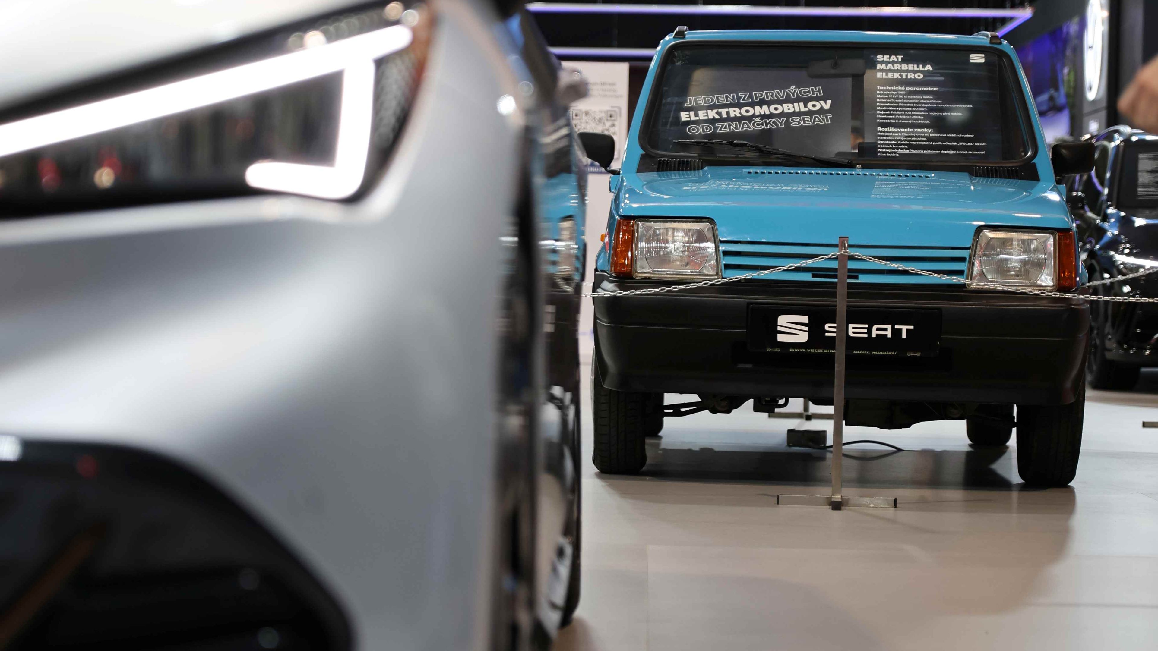 Dve autá sú vystavené na autosalóne. Predné auto je staršej modrej farby a zadné je moderné striebornej farby. Na pozadí je logo automobilky Volkswagen.