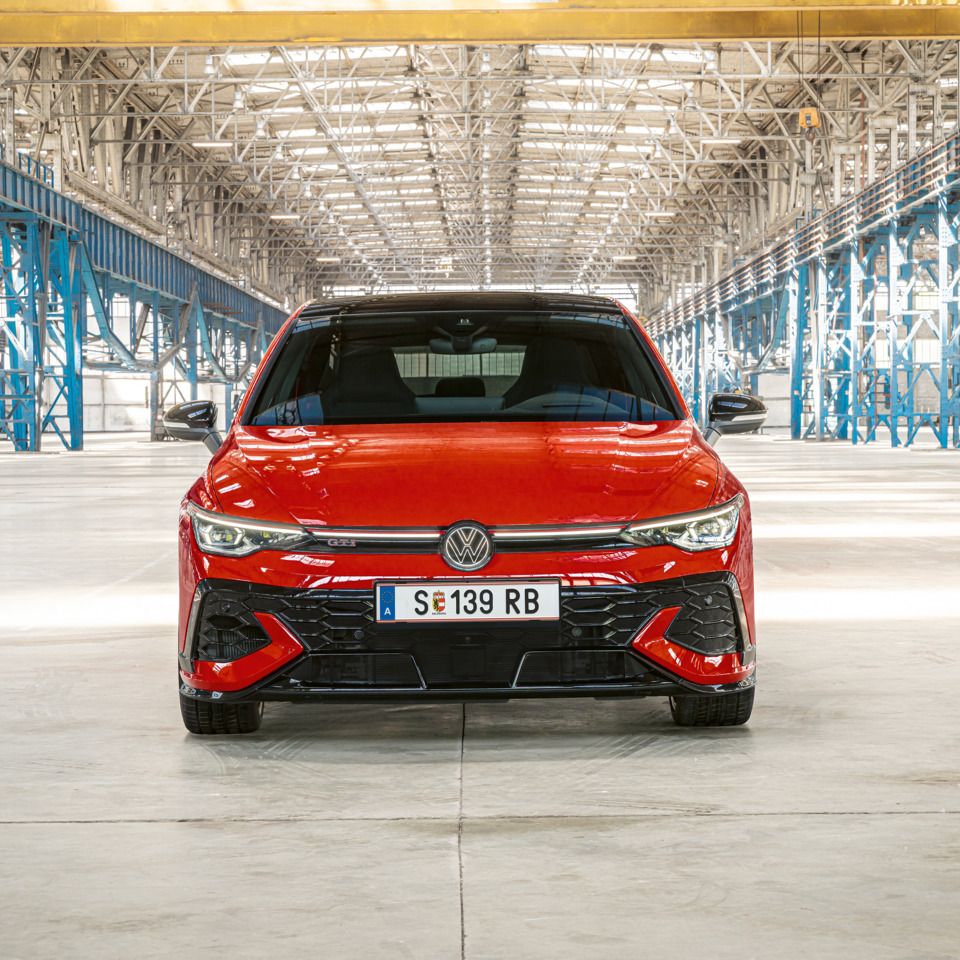 Červený Golf GTI je zaparkovaný vo veľkom, jasne osvetlenom sklade. Auto je zobrazené spredu. V pozadí sú viditeľné kovové konštrukcie a stĺpy.