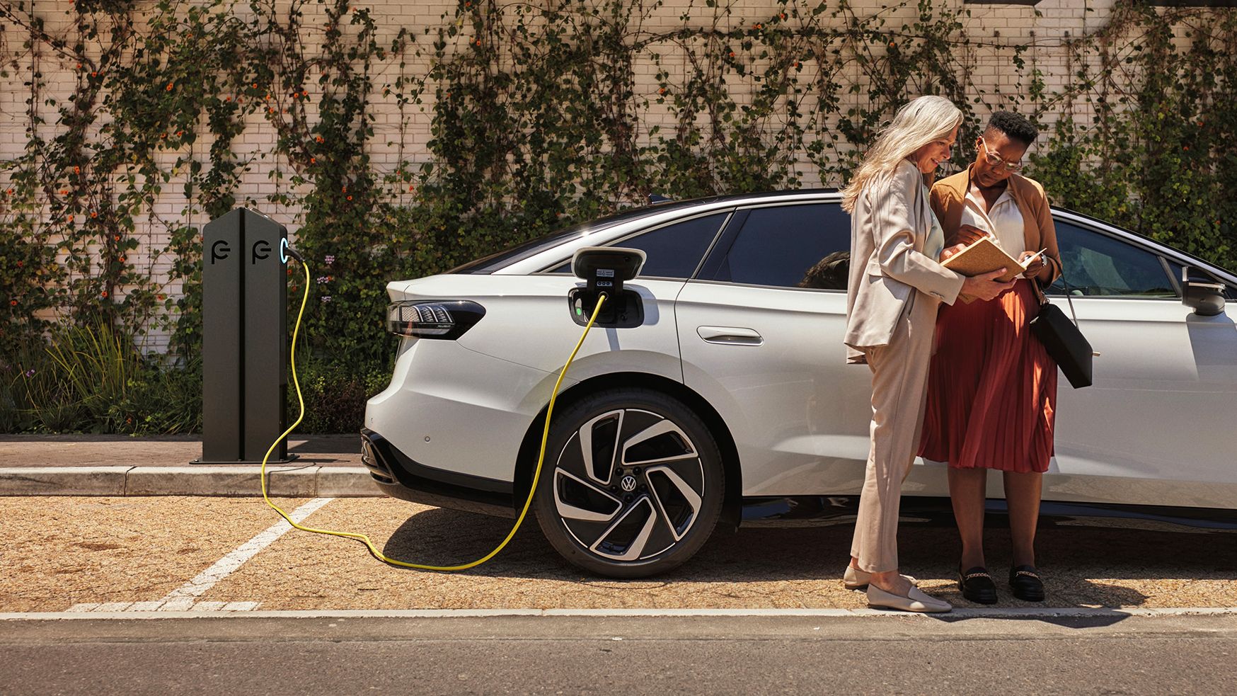 Dve ženy stoja s knihou v ruke vedľa bieleho VW ID., ktorý sa nabíja pri verejnej nabíjacej stanici.