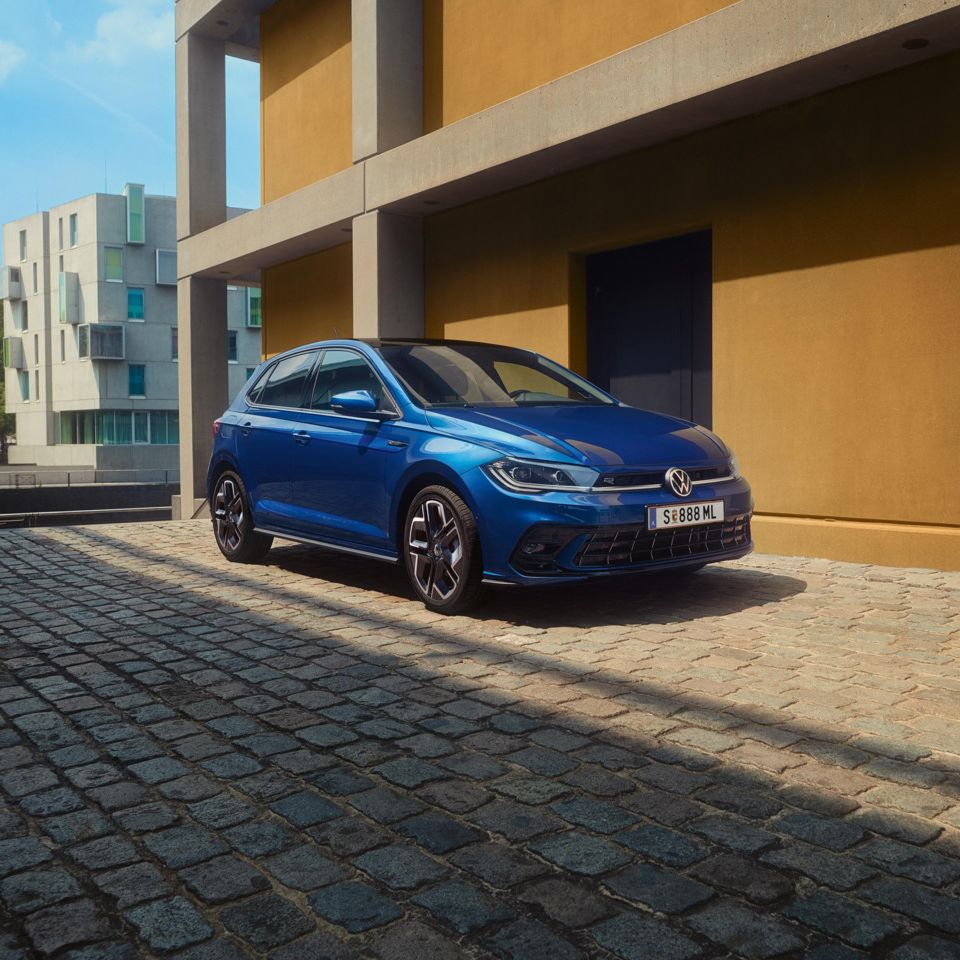 Modré Polo Volkswagen zaparkované na dláždenej ceste vedľa moderných bytových domov. V pozadí vidieť zeleň a zrejme cestu.