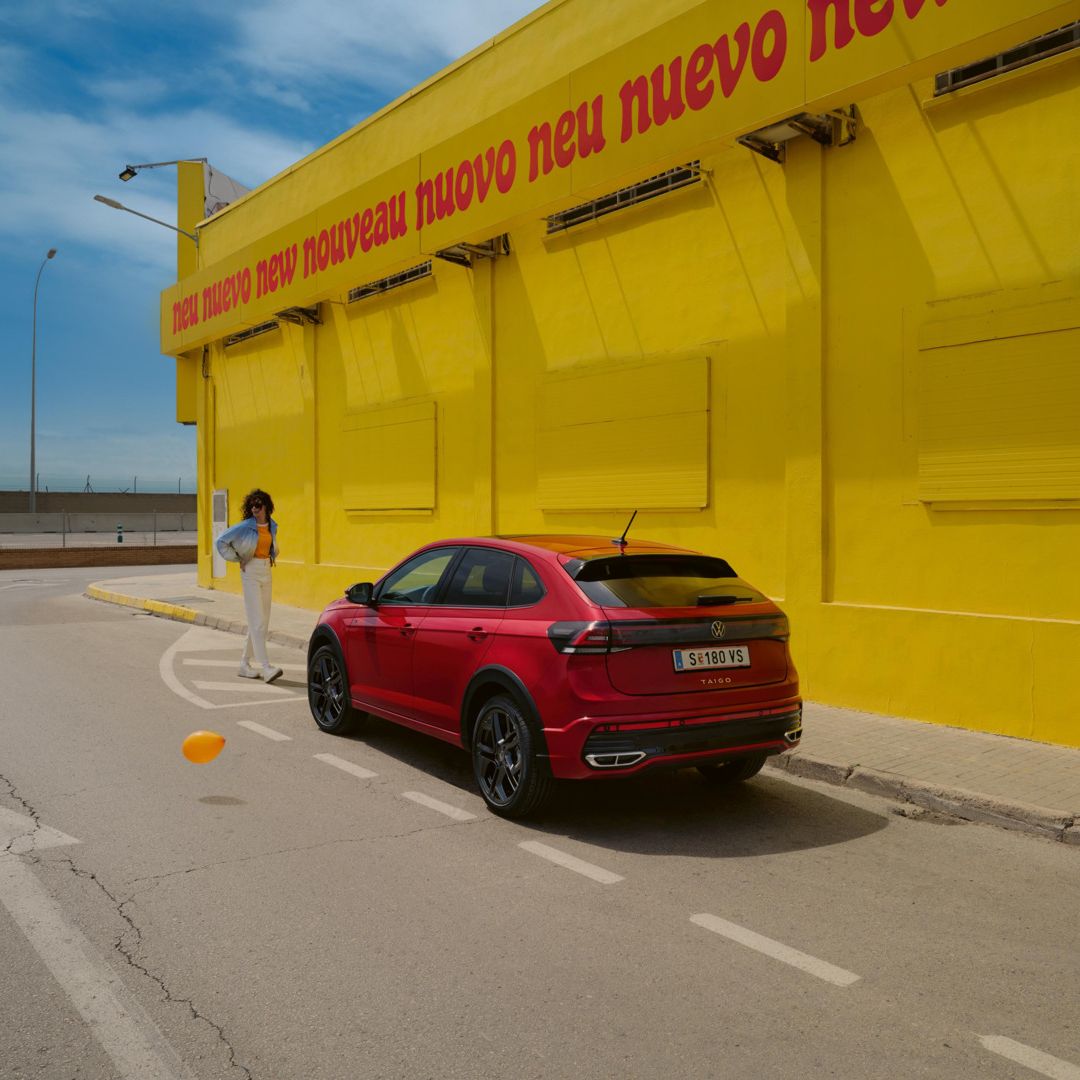 Červené Taigo R-Line je zaparkované pred žltou budovou s nápisom "neuvo neu nouveau neu nuevo neu". K vozidlu sa popri stene približuje mladá žena v modrej bunde. Vozidlo stojí na parkovacom mieste pri ceste.