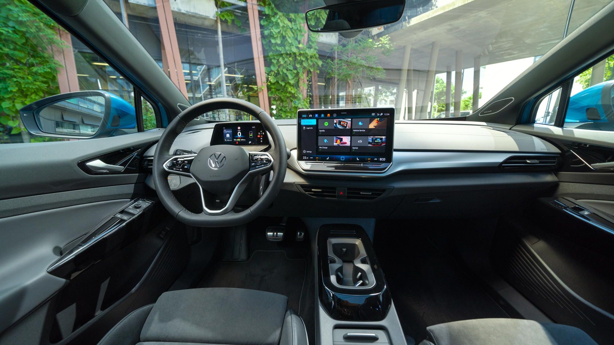 Interiér moderného Volkswagen ID.4. Na obrázku je elegantná palubná doska s integrovaným dotykovým displejom. Táto obrazovka zobrazuje rôzne aplikácie a informácie. Vidieť tiež časť predných sedadiel, digitálny prístrojový panel, stredovú konzolu a obklad dverí v modernom dizajne. Cez čelné sklo vidieť pohľad na modernú stavbu.