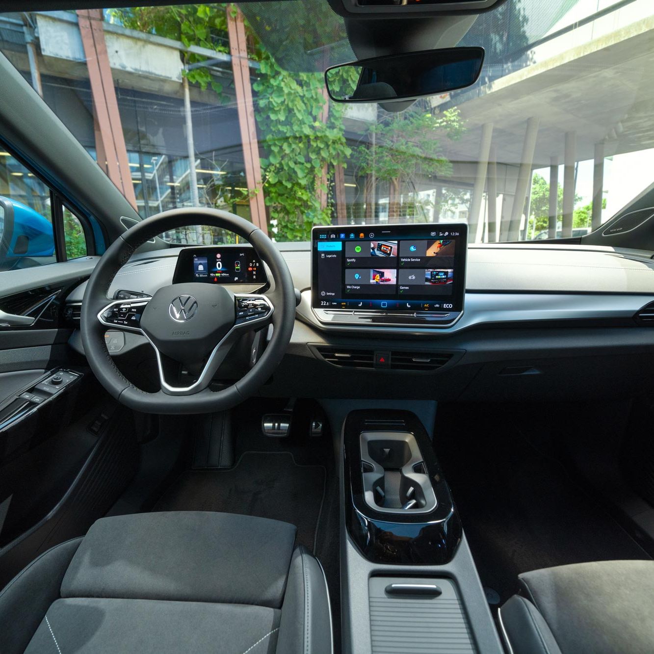 Interiér moderného Volkswagen ID.4. Na obrázku je elegantná palubná doska s integrovaným dotykovým displejom. Táto obrazovka zobrazuje rôzne aplikácie a informácie. Vidieť tiež časť predných sedadiel, digitálny prístrojový panel, stredovú konzolu a obklad dverí v modernom dizajne. Cez čelné sklo vidieť pohľad na modernú stavbu.