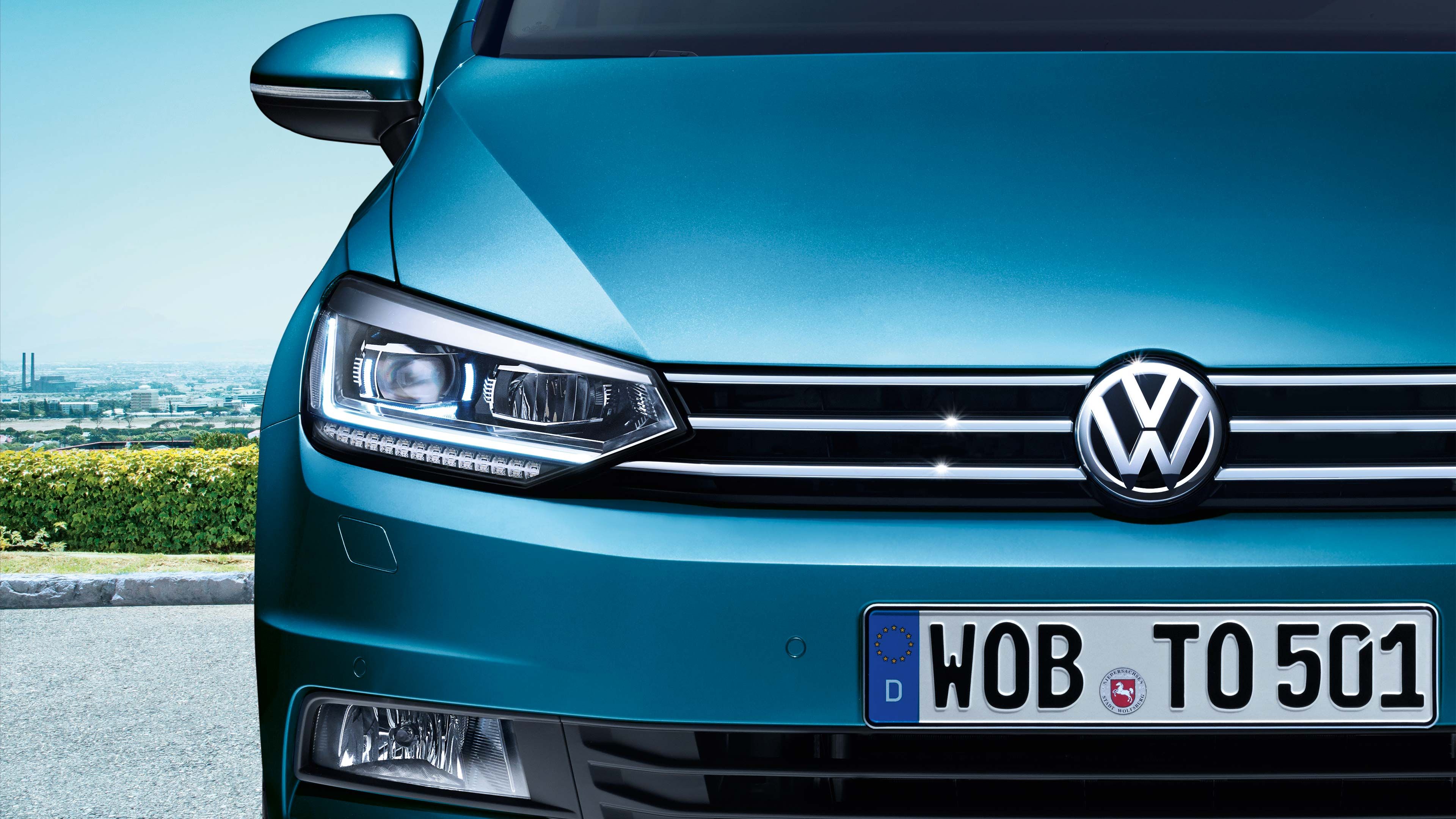 Automobil VW z predného pohľadu s nemeckou registračnou značkou. Karoséria je modrej farby. V pozadí vidieť zeleň a mesto.