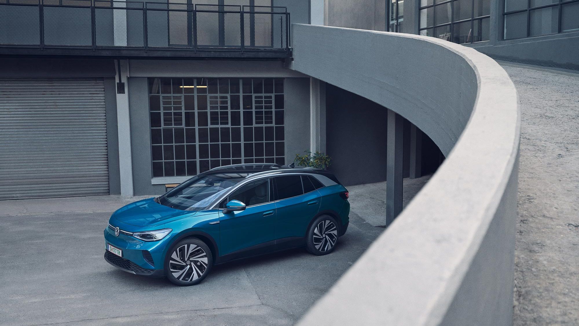 Modré SUV je zaparkované na betónovej ploche vedľa moderných budov. Auto má štýlový dizajn. Scéna je pokojná bez prítomnosti ľudí.