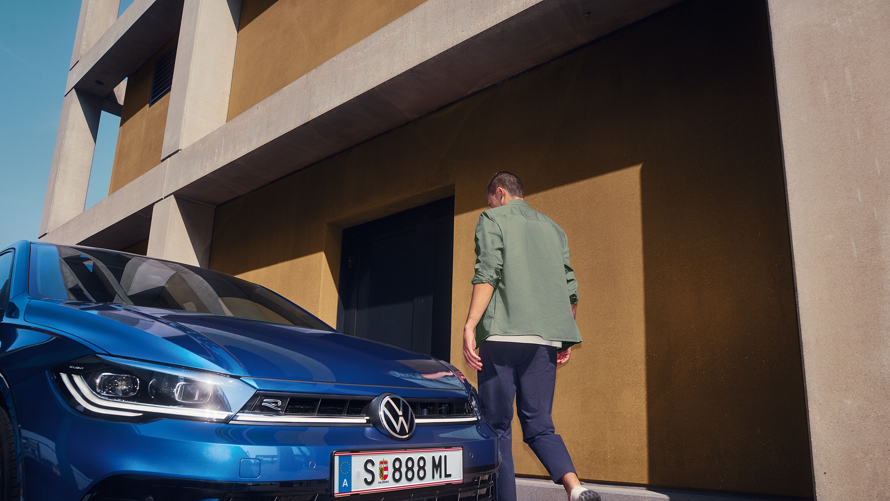 Muž kráča k budove, vedľa nej je zaparkované modré auto značky Volkswagen. Teplé farby budovy a modrá farba auta vzájomne kontrastujú. Muž má na sebe zelenú bundu a tmavé nohavice.