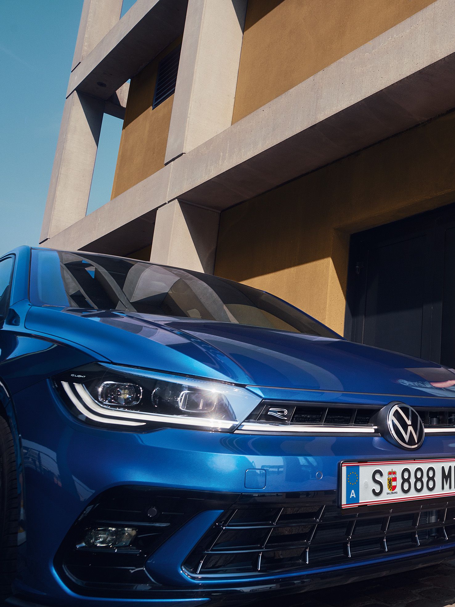 Muž kráča k budove, vedľa nej je zaparkované modré auto značky Volkswagen. Teplé farby budovy a modrá farba auta vzájomne kontrastujú. Muž má na sebe zelenú bundu a tmavé nohavice.