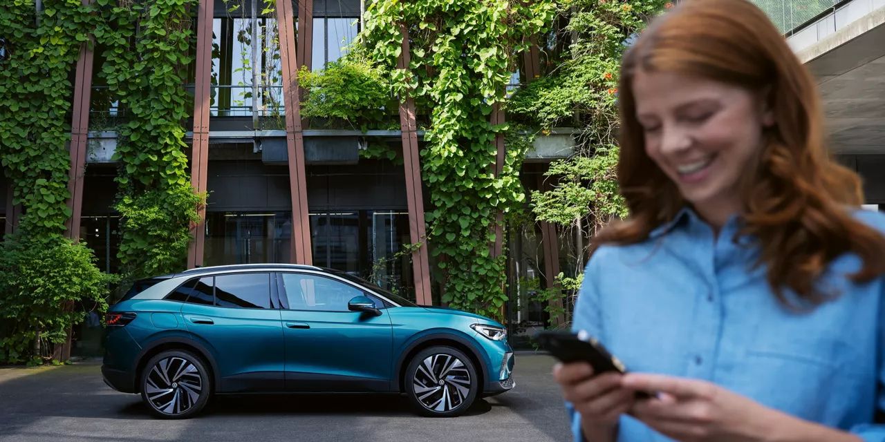 Modrý elektromobil zaparkovaný pred budovou obrastenou zeleňou. Nezaostrený pohľad na ženu v popredí s ryšavými vlasmi, ktorá drží mobilný telefón a pozerá sa na neho.