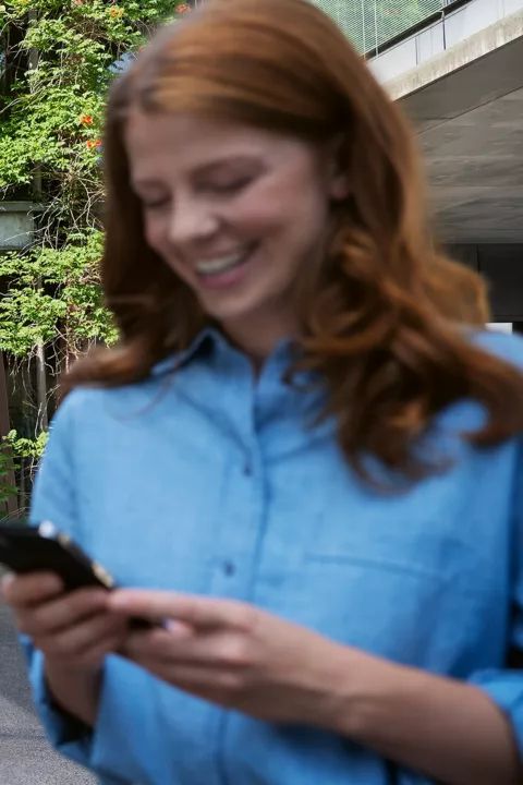 Modrý elektromobil zaparkovaný pred budovou obrastenou zeleňou. Nezaostrený pohľad na ženu v popredí s ryšavými vlasmi, ktorá drží mobilný telefón a pozerá sa na neho.