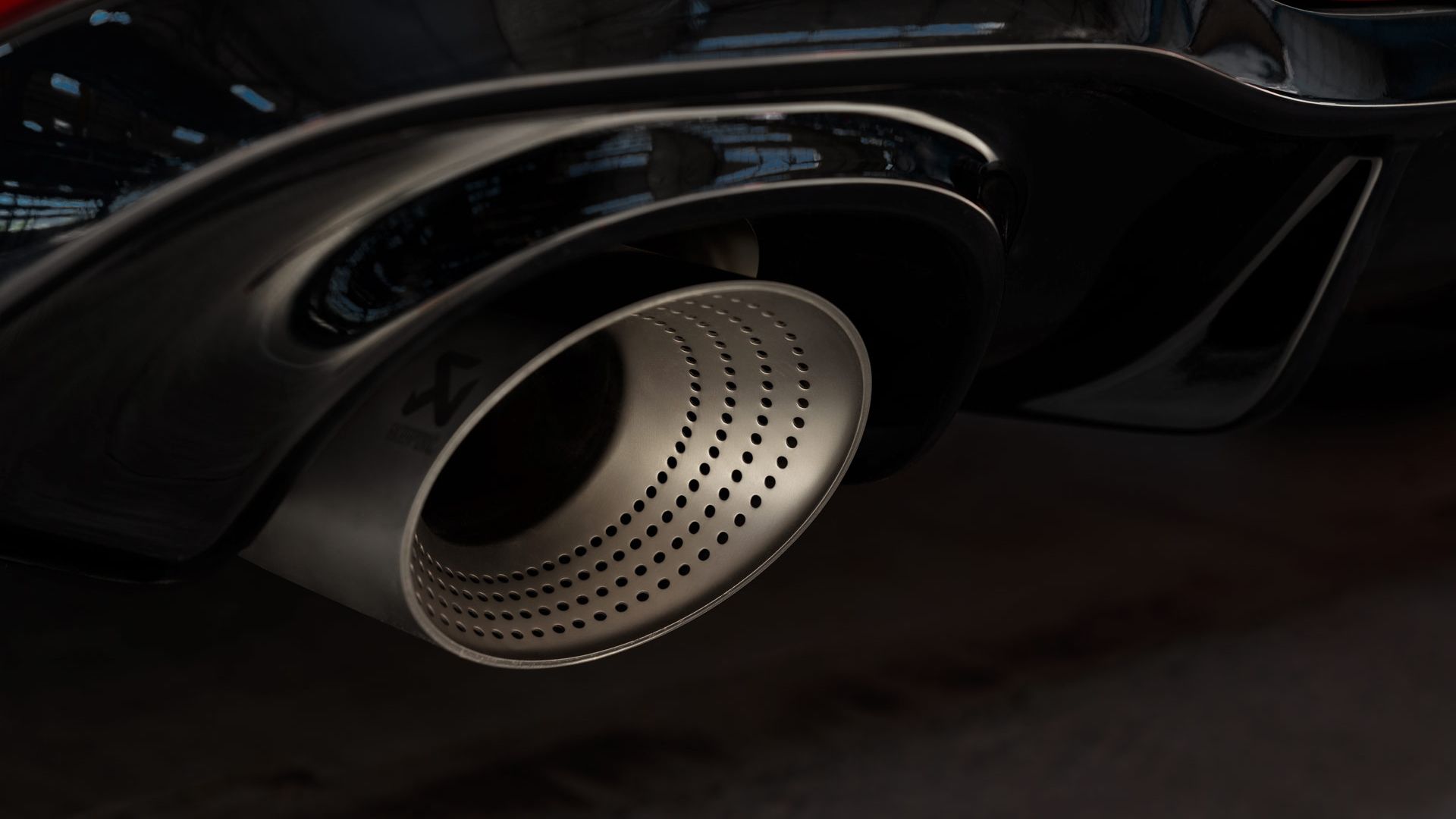 Detailný záber na výfuk športového auta so striebornou perforovanou koncovkou. Výfuková rúra patrí červenému modelu Golf GTI a lesklá čierna karoséria je ľahko rozpoznateľná. V pozadí je vidieť rozmazaný obrys budovy.