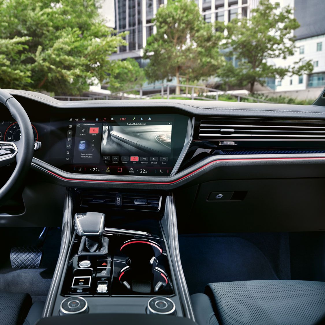Pohľad do interiéru VW Touareg Elegance eHybrid, na displeji je zobrazené nastavenie jazdného režimu. Cez predné okno vidieť mestské prostredie a zeleň.