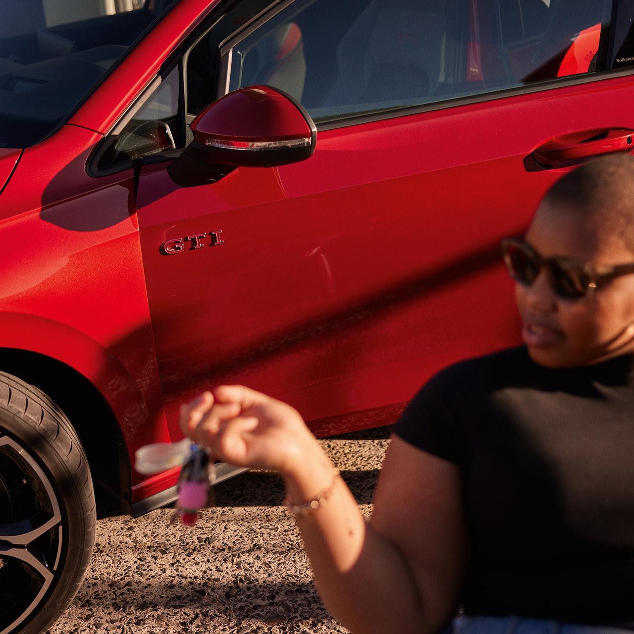 Detailný záber na predné partie červeného VW Golf GTI zboku so ženou v popredí, ktorá sedí na stoličke pred automobilom. Muž za ňou stojí v otvorených dverách vodiča. Vozidlo stojí na štrkovej ceste.
