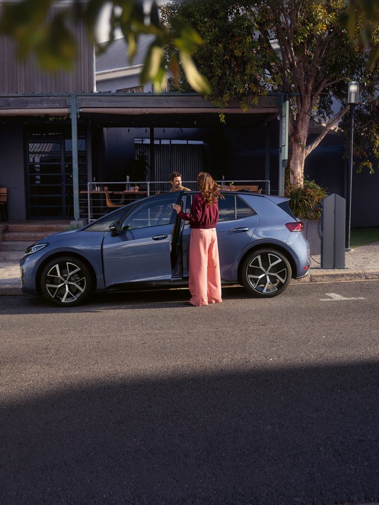 Žena a muž nastupujú do modrého VW ID.3 Neo, ktorý parkuje pred kaviarňou.