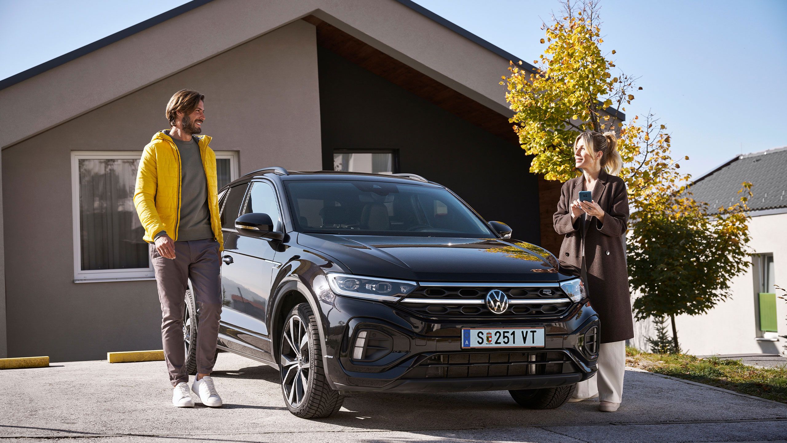 Muž a žena stoja so smartfónom pred vozidlom Volkswagen pri rodinnom dome.